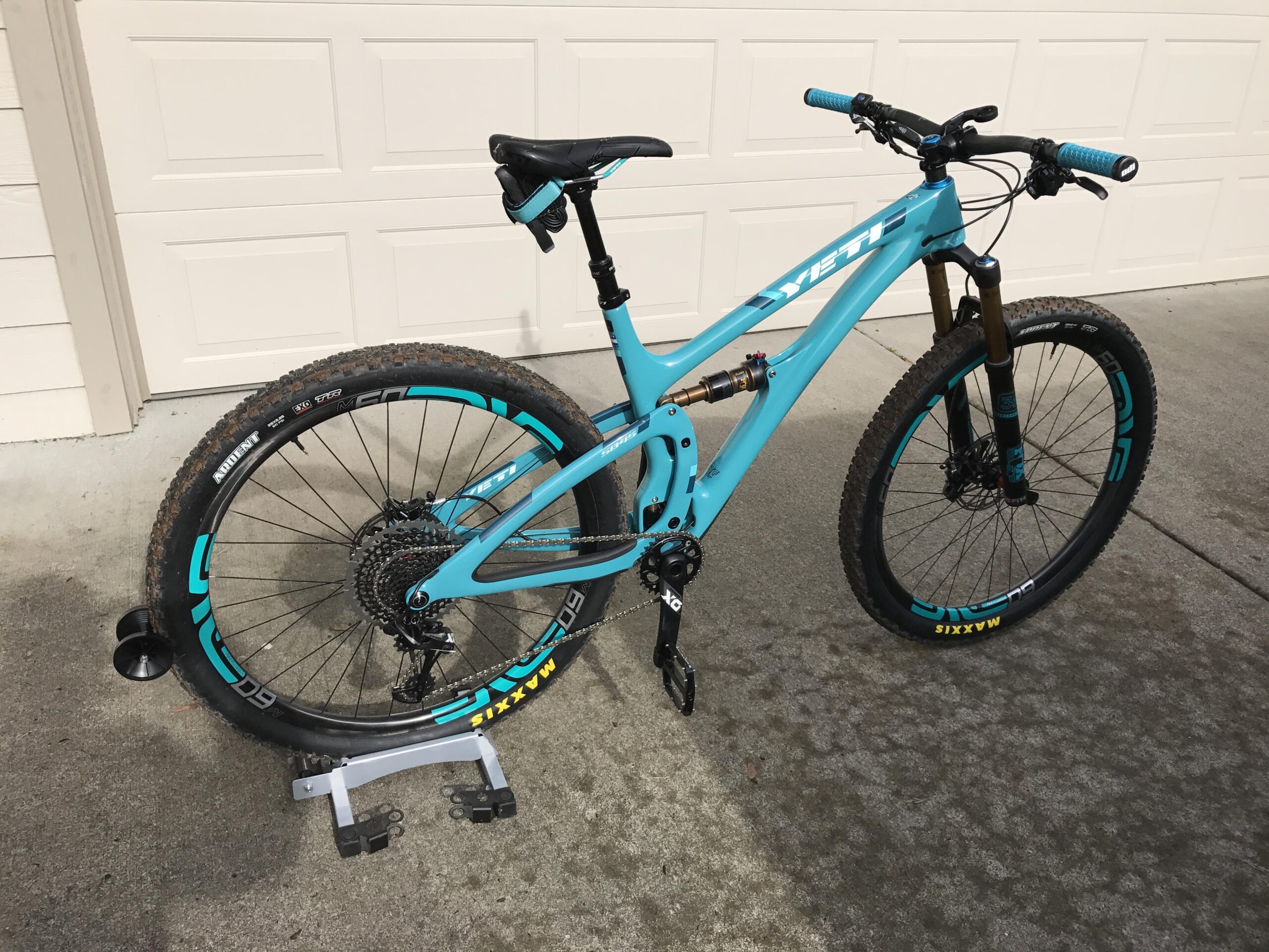 Yeti SB4.5c: A bright turquoise mountain bike with black and teal accents is shown positioned on a bike stand in front of a beige garage door. The bike features thick tires designed for off-road terrain, a sturdy frame with a suspension system, and textured handlebar grips. The background consists of a smooth concrete floor, emphasizing the bike's details.