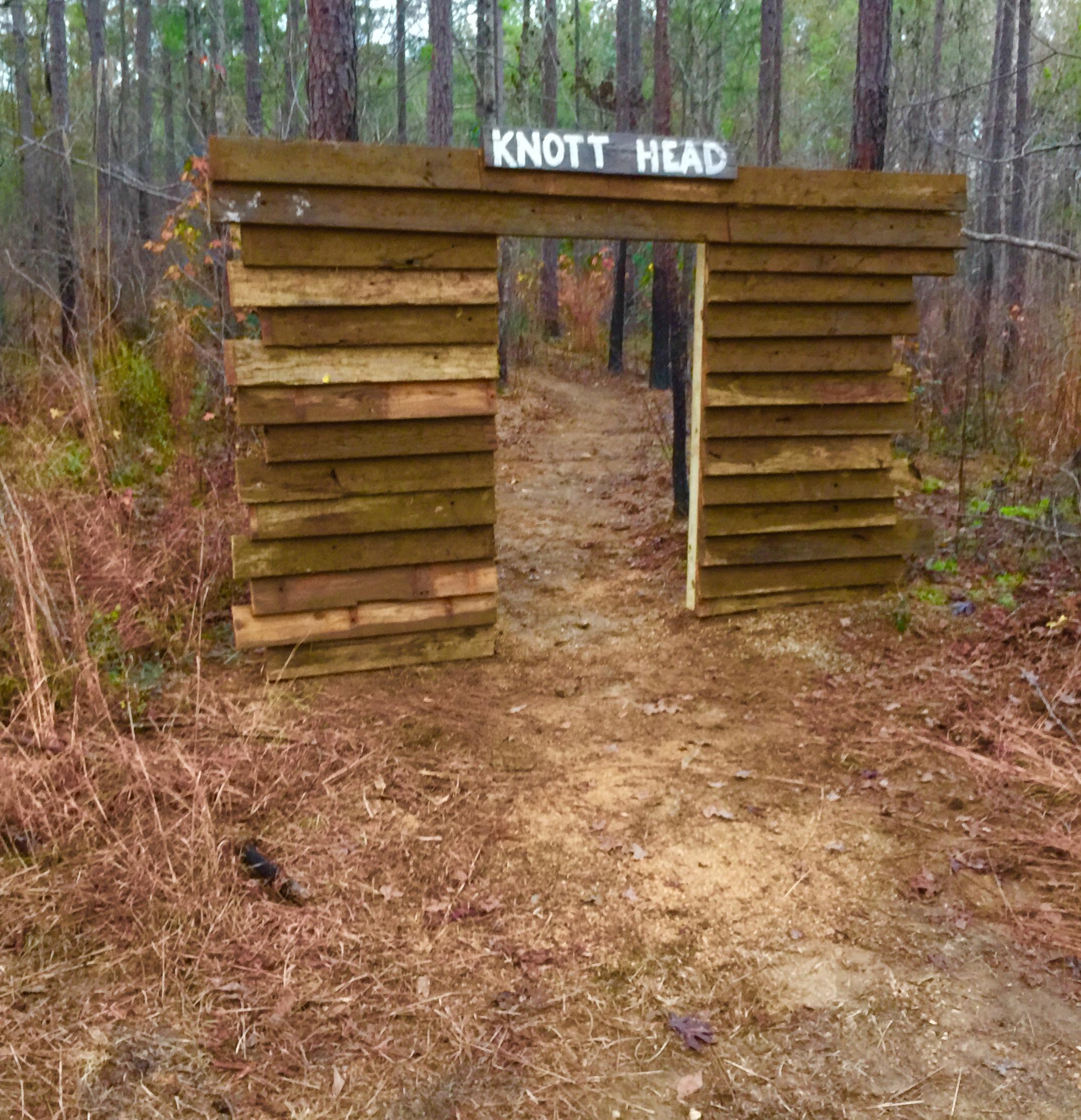 A wooden archway labeled "KNOTT HEAD" at the entrance of a forest trail surrounded by tall pine trees and sparse underbrush. The path is dirt, with scattered leaves and small twigs visible on the ground.