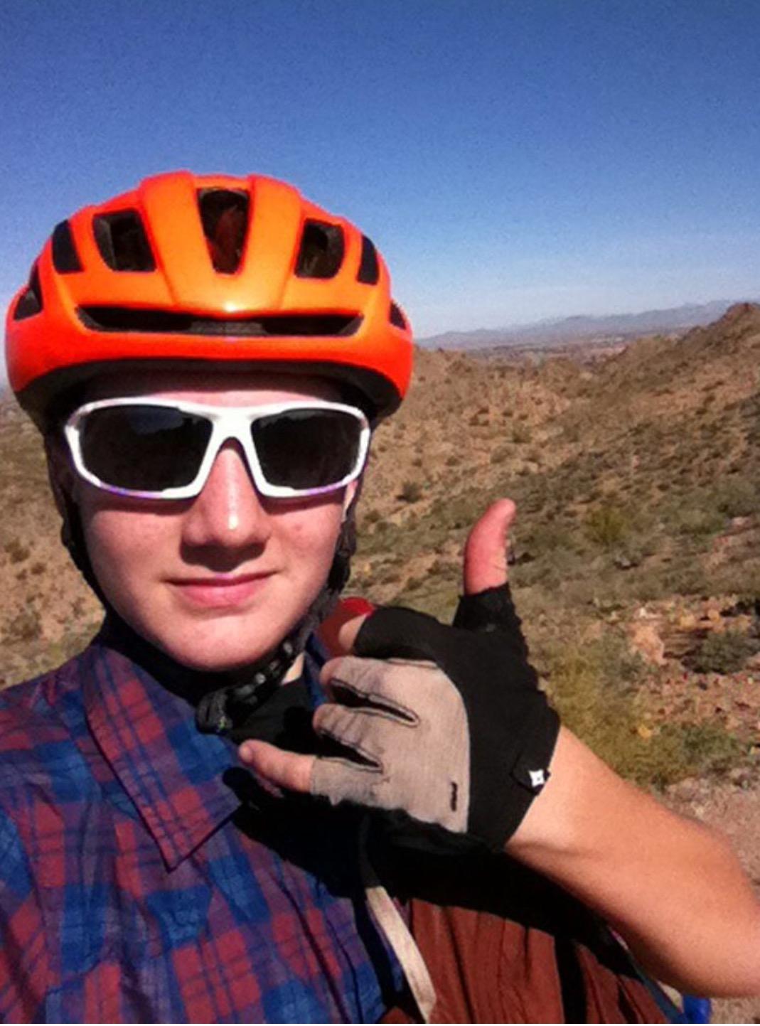 A young person wearing an orange biking helmet and sunglasses gives a thumbs-up gesture while taking a selfie on a sunny day. They are dressed in a plaid shirt and fingerless gloves, with a mountainous landscape visible in the background. San Tan mountain bike trail.