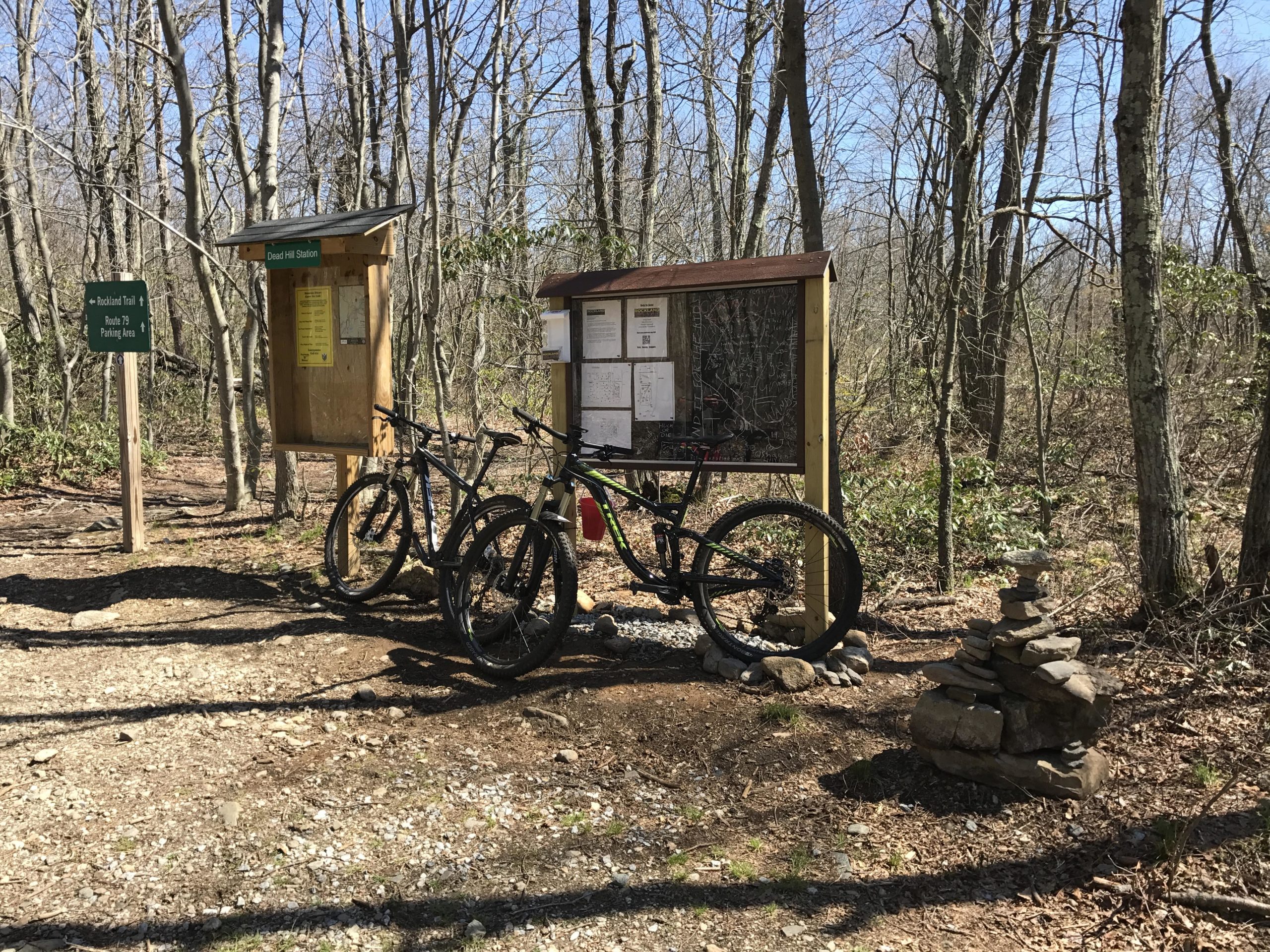 Two mountain bikes are parked next to a wooden information board and a signpost in a forested area. The board features trail maps and notices, while the sign indicates directions for nearby trails. A small stack of stones is placed on the ground beside the bikes. The scene is set in a sunny, wooded environment with bare trees. Rockland Preserve mountain bike trail.