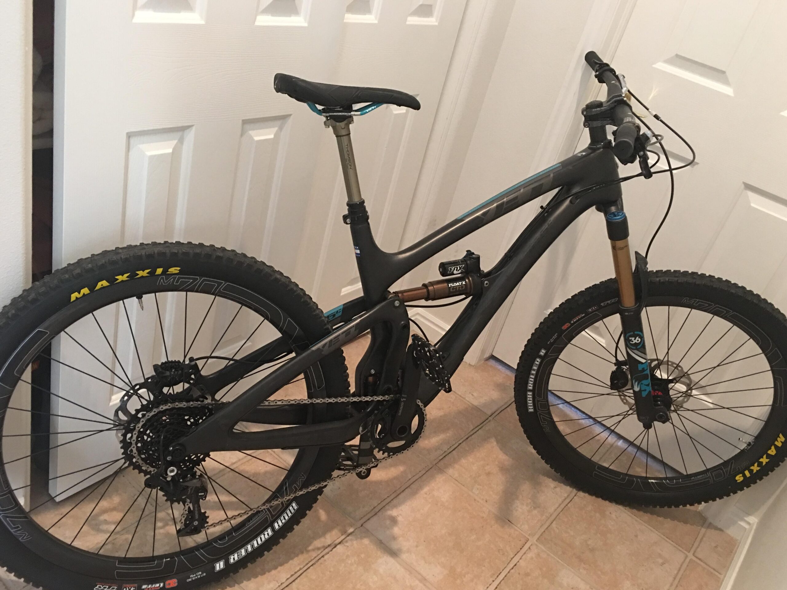 Yeti SB6: A black mountain bike with blue and gold accents displayed in an indoor setting, positioned against white doors. The bike features thick tires marked "MAXXIS," a suspension fork, and a geared drivetrain. The floor is tiled.