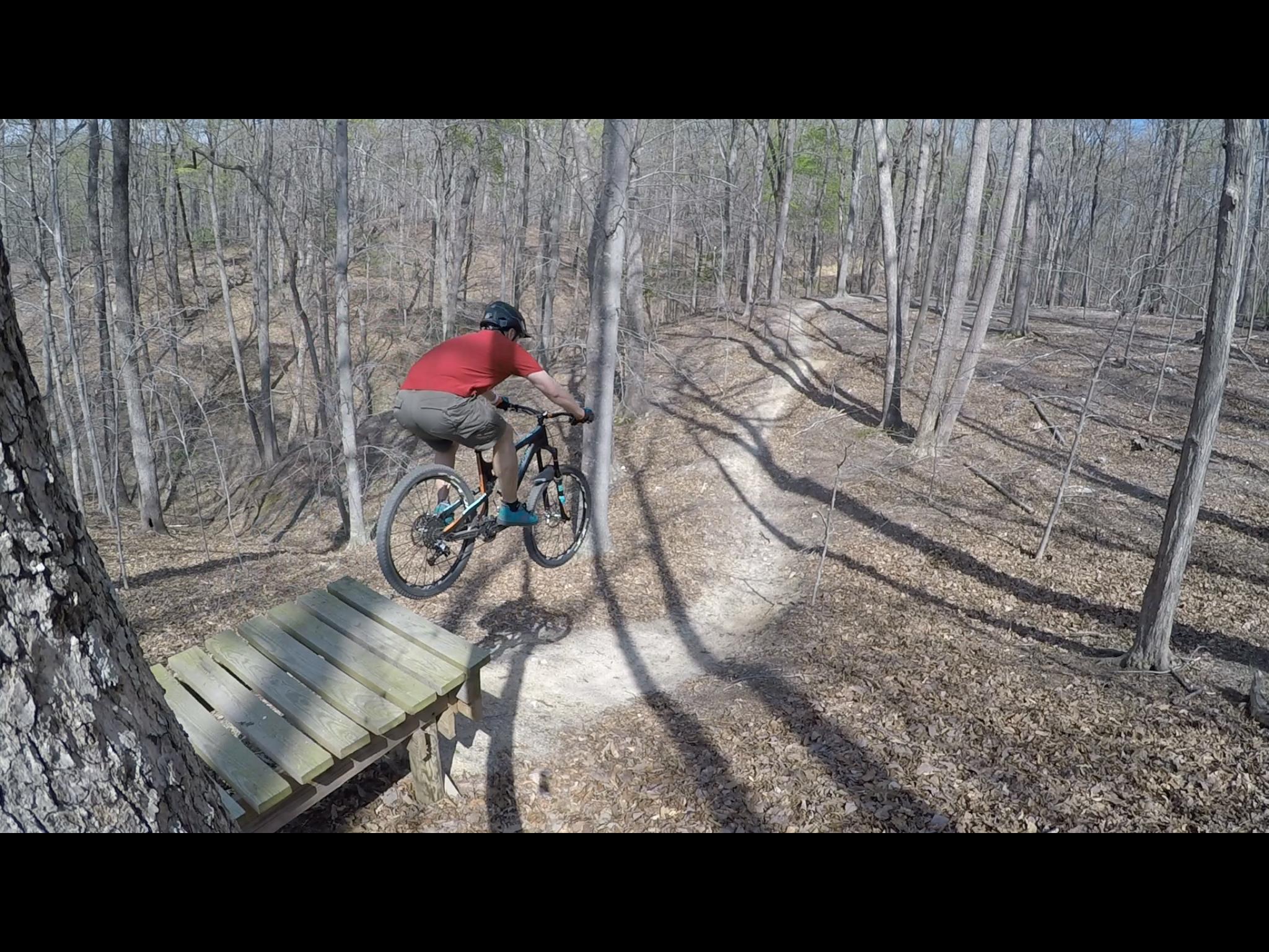 A cyclist in a red shirt and helmet jumps off a wooden ramp in a wooded area, mid-air above a dirt trail surrounded by bare trees and fallen leaves. Fountainhead Regional Park mountain bike trail.