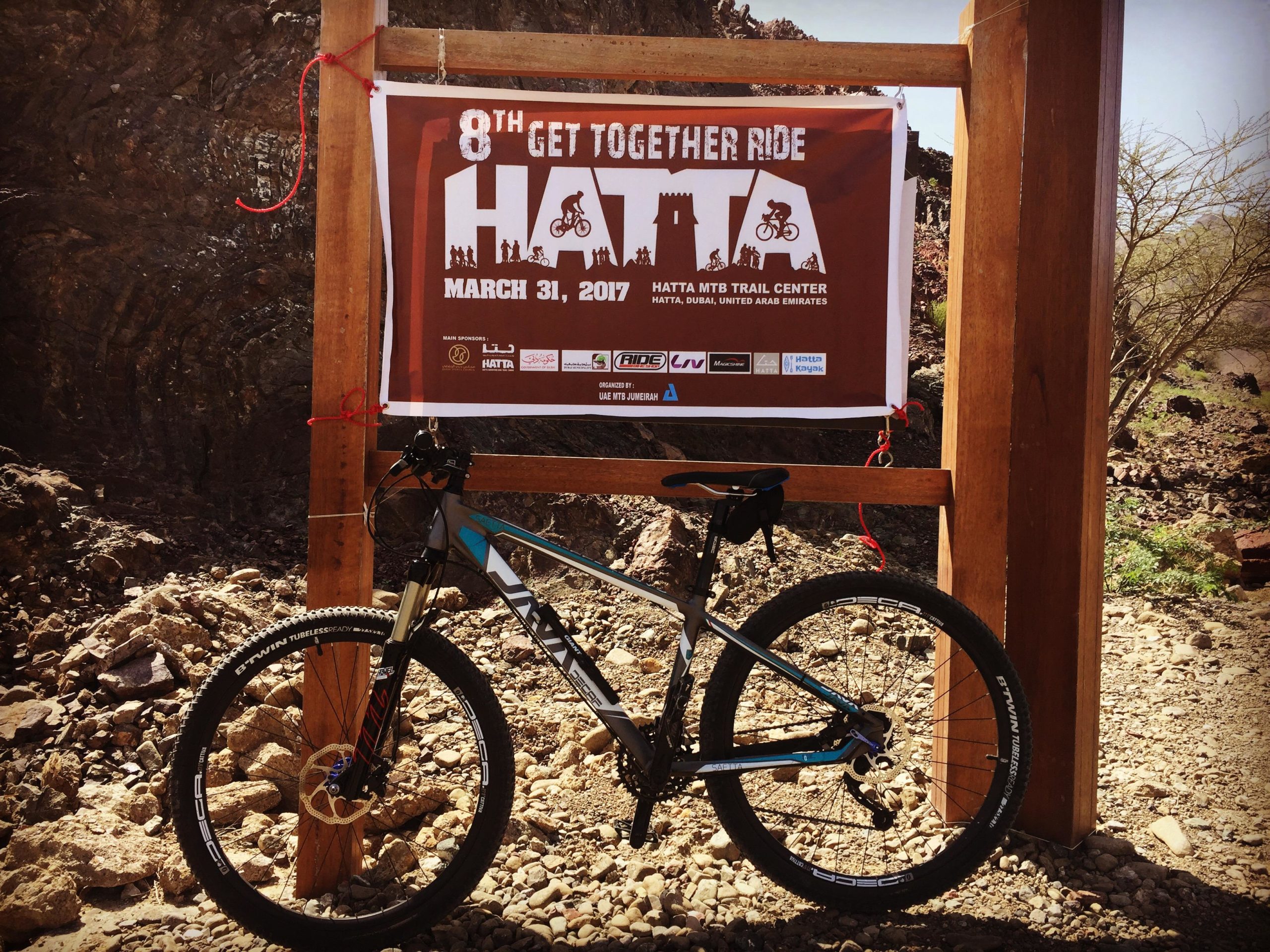 A mountain bike is parked next to a wooden sign announcing the "8th Get Together Ride" at Hatta MTB Trail Center, dated March 31, 2017. The sign features graphics of cyclists and includes sponsor logos, set against a rocky landscape. Showka Trails mountain bike trail.