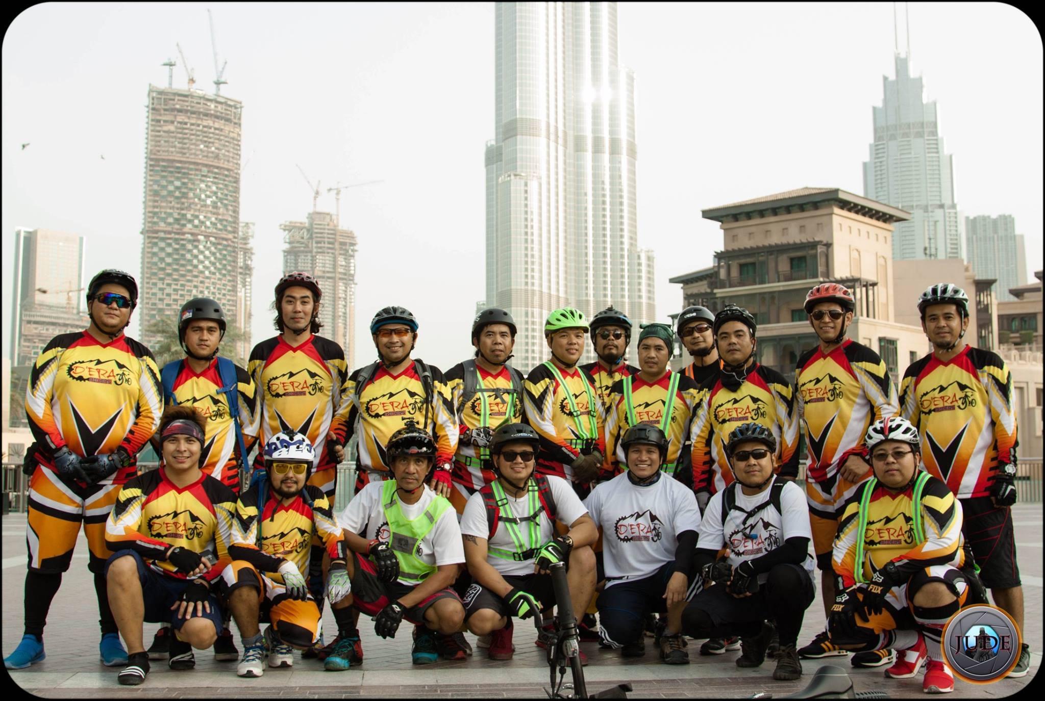 A diverse group of cyclists poses for a photo in colorful cycling jerseys, showcasing vibrant designs in red, yellow, and black. The group stands in front of a skyline featuring tall buildings, including a notable skyscraper, with construction cranes visible in the background. The cyclists include individuals wearing helmets and some holding bicycles, emphasizing a sense of community and passion for cycling. Showka Trails mountain bike trail.
