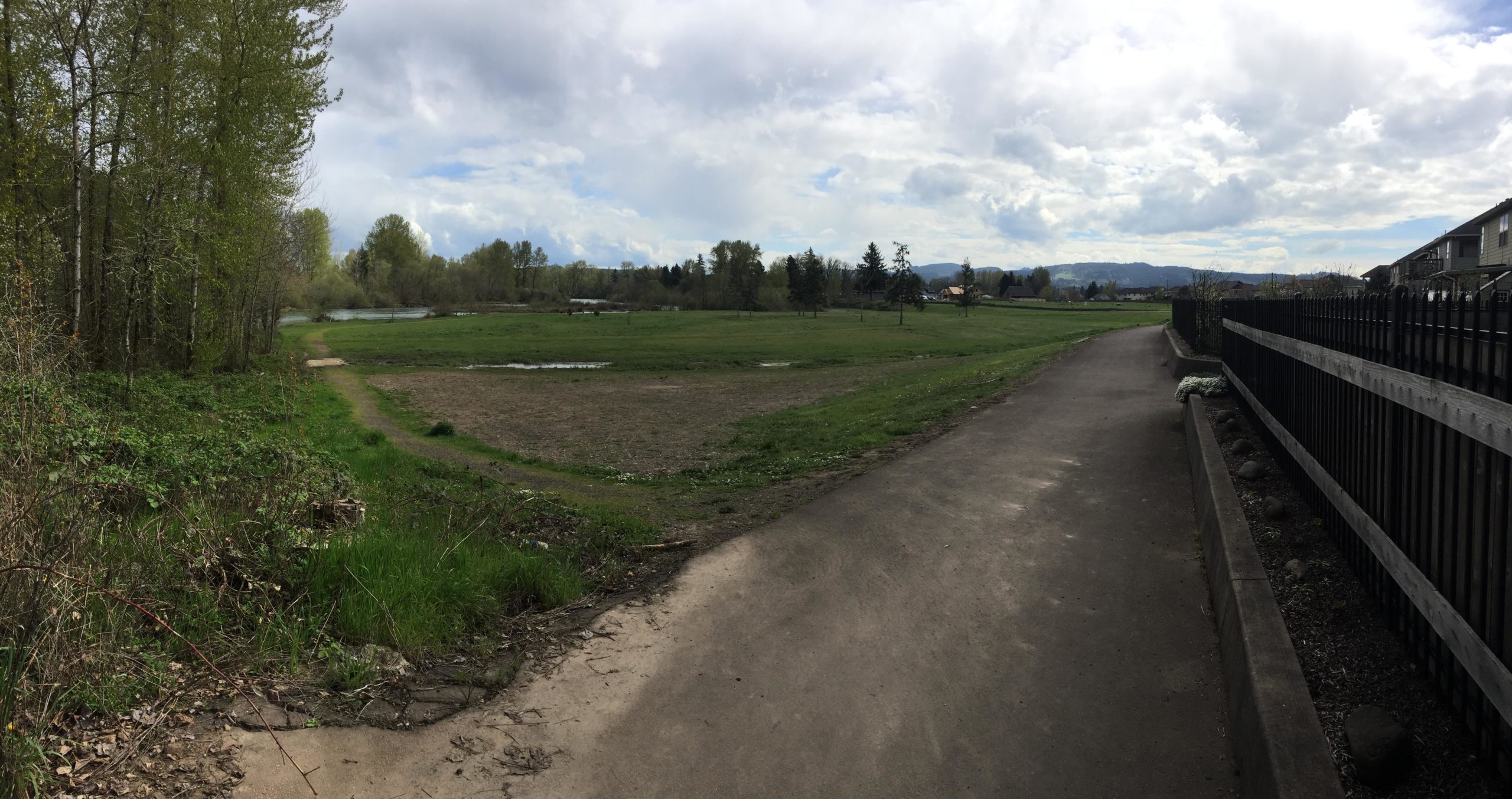 A panoramic view of a lush green field bordered by a dirt path and a wooden fence. The scene includes trees in the background, a calm waterway, and a partly cloudy sky, capturing a serene outdoor setting. Lebanon Dikes mountain bike trail.