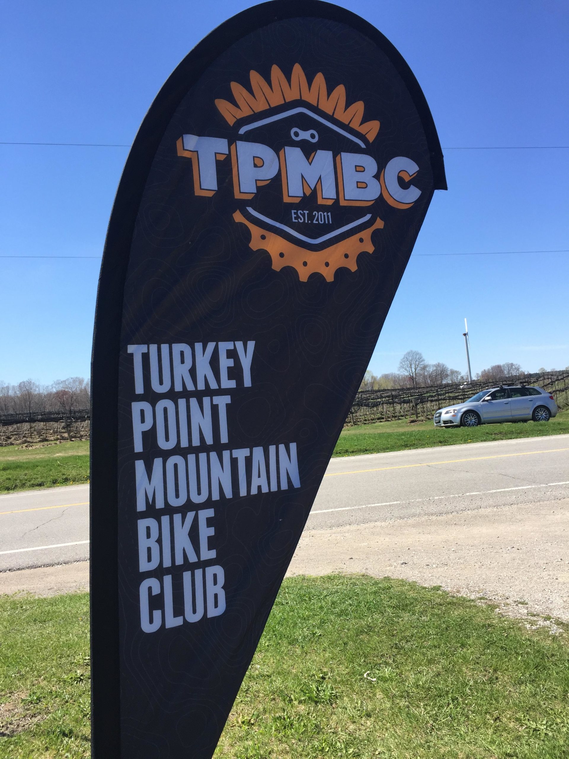 Flag for the Turkey Point Mountain Bike Club (TPMBC), featuring the club's logo and establishment year, set against a clear blue sky with a grassy area and road in the background. Turkey Point Provincial Park mountain bike trail.