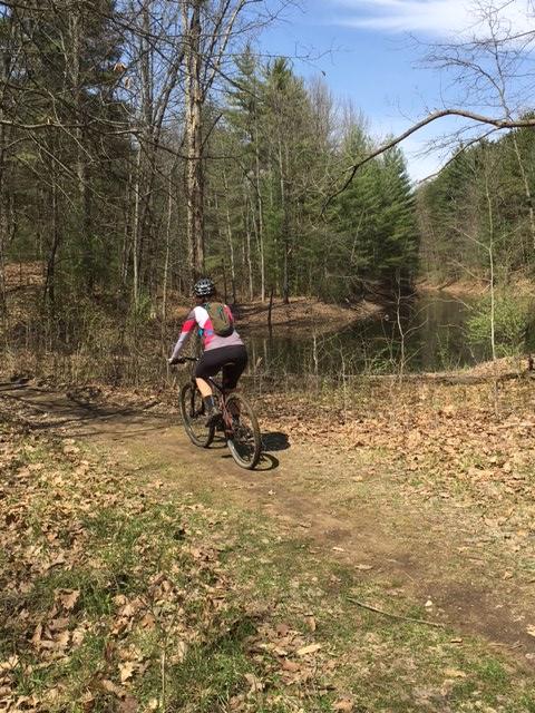 A person riding a mountain bike on a dirt trail surrounded by trees, with a small pond visible in the background. The scene is set in a wooded area during a sunny day, with some patches of green grass and fallen leaves on the ground. Advanced Access Trail mountain bike trail.