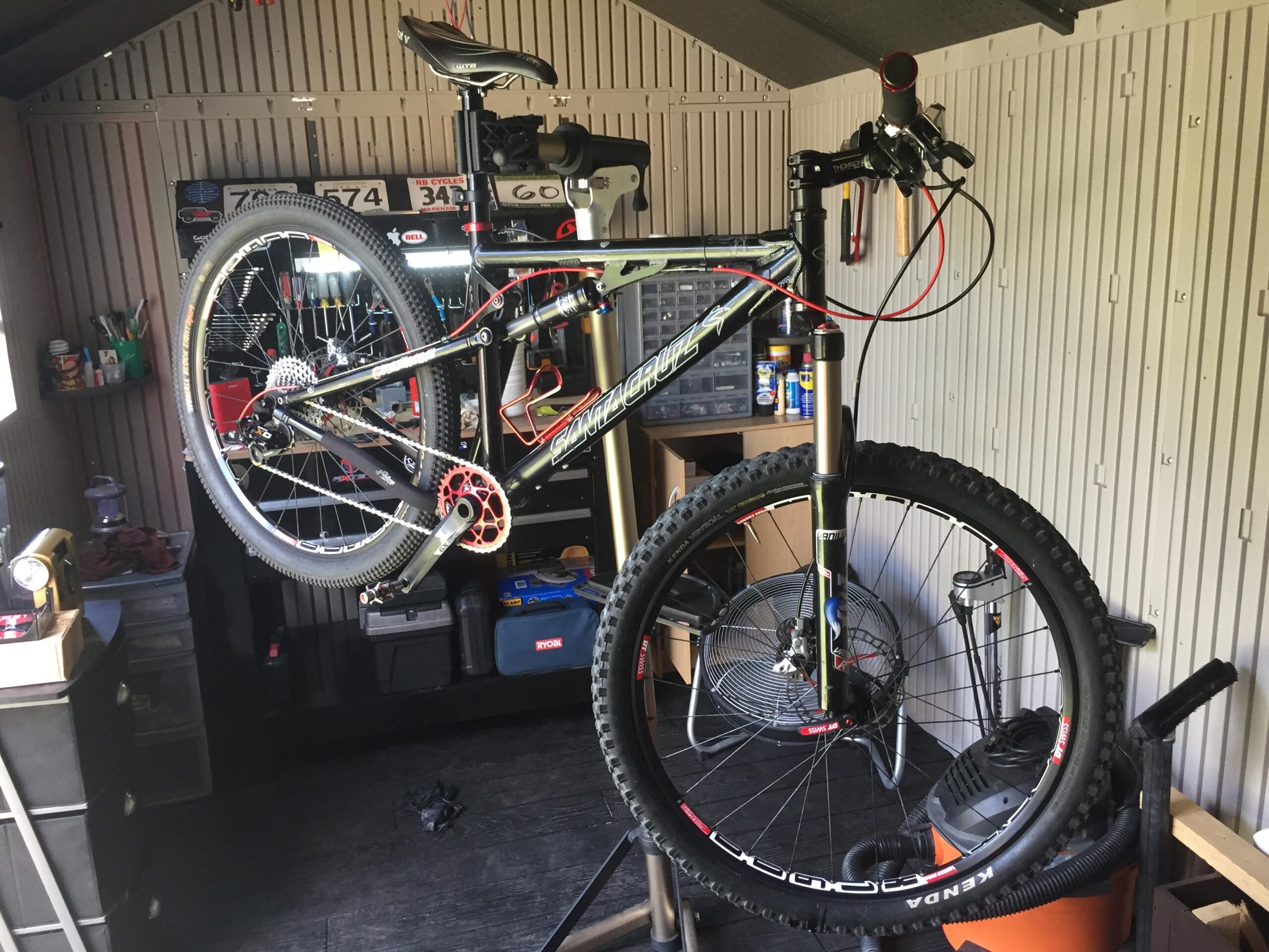 Santa Cruz Blur: A mountain bike is mounted on a repair stand in a workshop, with various tools and equipment visible in the background. The bike features a black and red frame, a prominent front suspension fork, and thick, knobby tires. The workspace shows a mix of cycling gear, tools, and storage, highlighting an environment dedicated to bike maintenance.