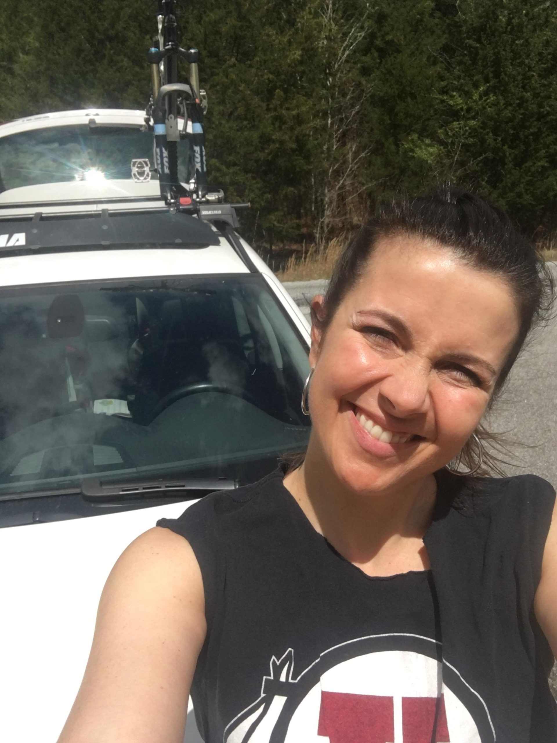 A smiling woman wearing a sleeveless black shirt stands in front of a white car with a bicycle mounted on the roof rack. Trees are visible in the background under bright sunlight. Enterprise South mountain bike trail.