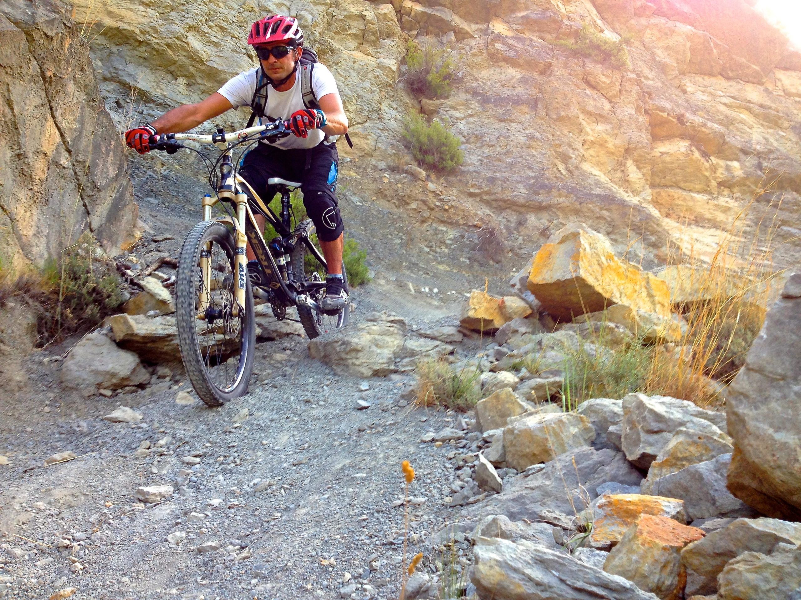 Mountain biker navigating a rocky trail, surrounded by rugged terrain and sunlight filtering through the rocks. The cyclist is wearing a helmet, sunglasses, and protective gloves, demonstrating an adventurous outdoor activity. MATEBA-RODECHE mountain bike trail.