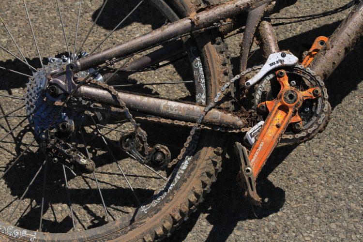 A close-up view of a muddy mountain bike's drivetrain, showcasing the rear cassette, chain, and crankset. The bike's frame is covered in dirt, emphasizing its use in off-road conditions. The orange crankset stands out against the grime, while the intricate designs on the tires are partially visible.