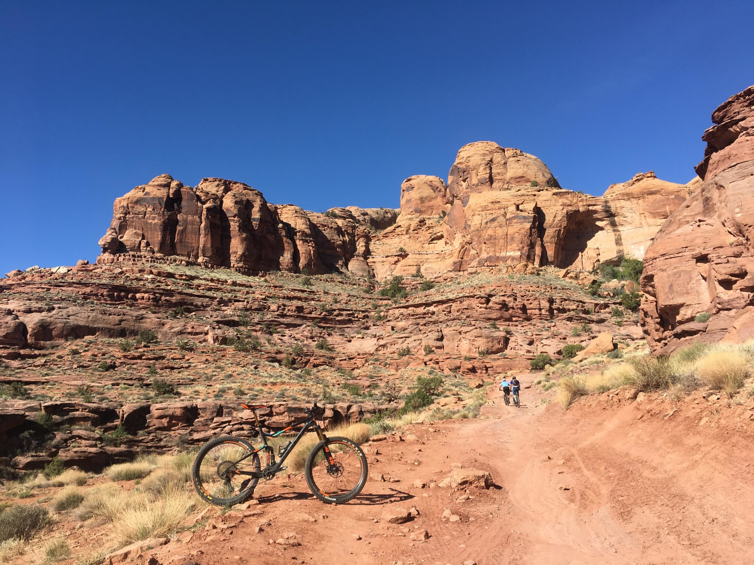 A mountain bike rests on a dirt trail in a rocky desert landscape, with steep, eroded cliffs in the background. Two hikers walk along the trail, surrounded by sparse vegetation under a clear blue sky. Captain Ahab mountain bike trail.