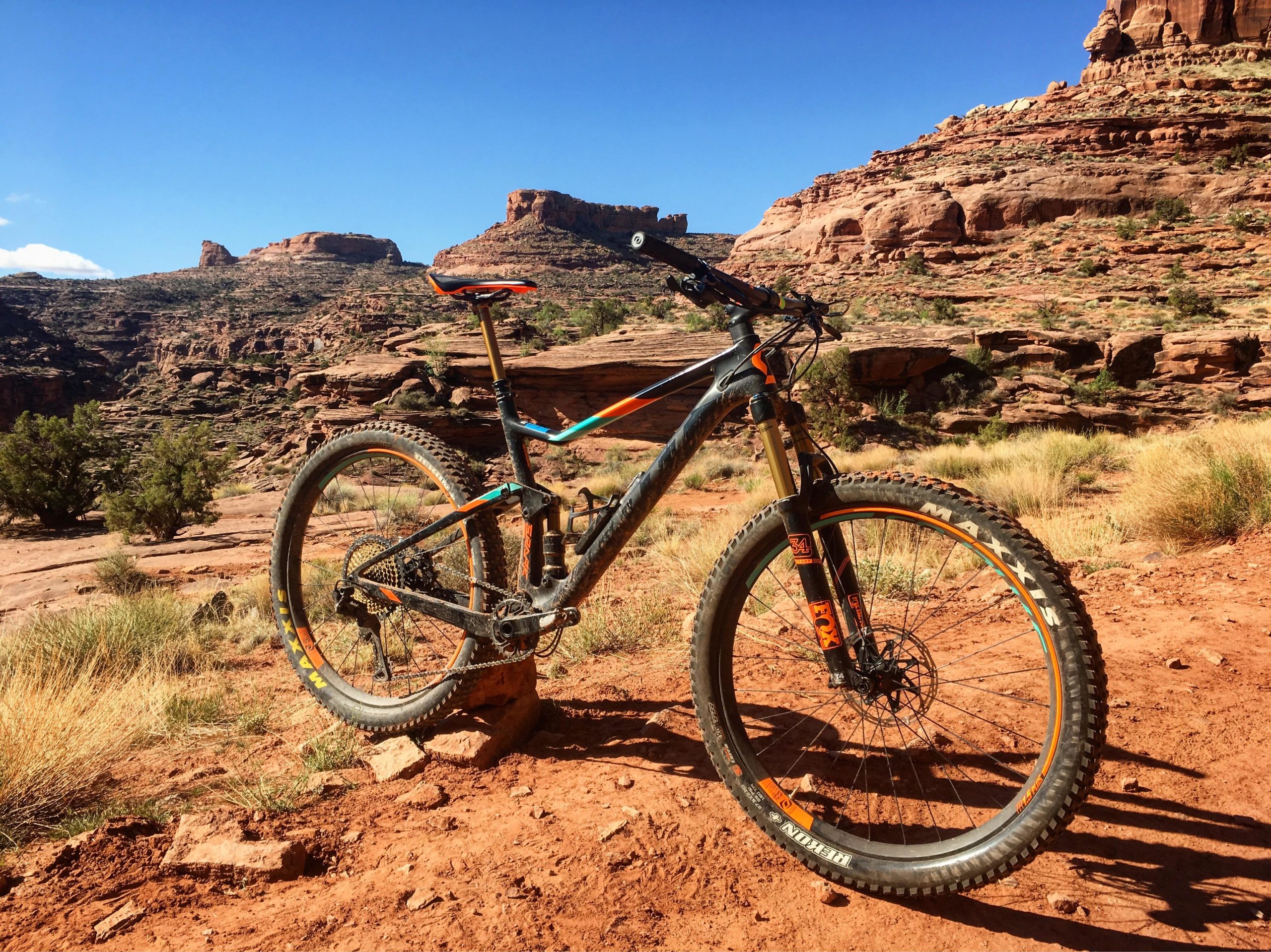 A mountain bike resting on rocky terrain in a desert landscape, with red soil and sparse vegetation. The background features rugged rock formations under a clear blue sky. Captain Ahab mountain bike trail.