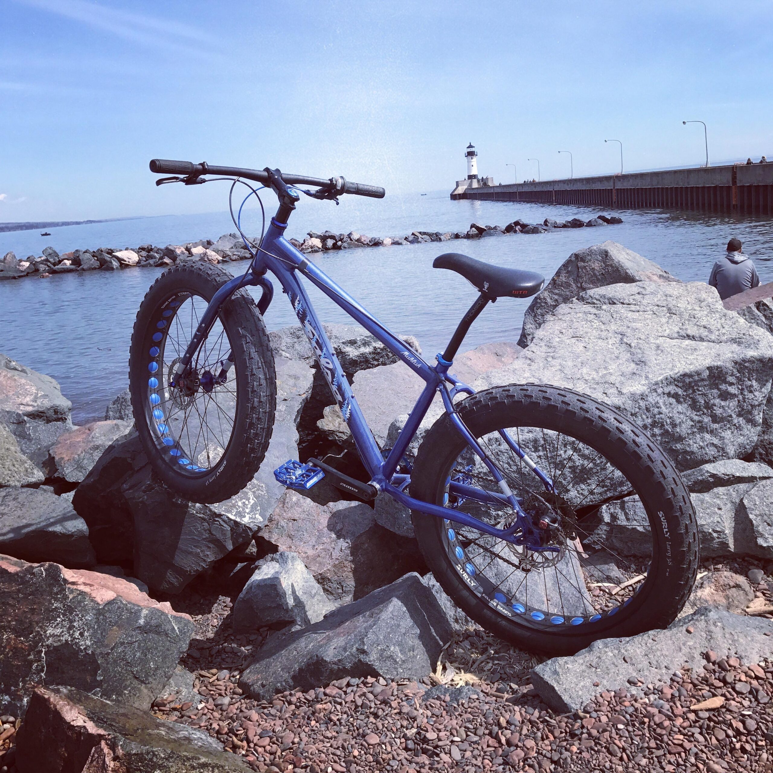 Salsa Mukluk 2: A blue fat tire bike resting on rocky shorelines near a body of water, with a lighthouse and pier visible in the background under a clear sky.