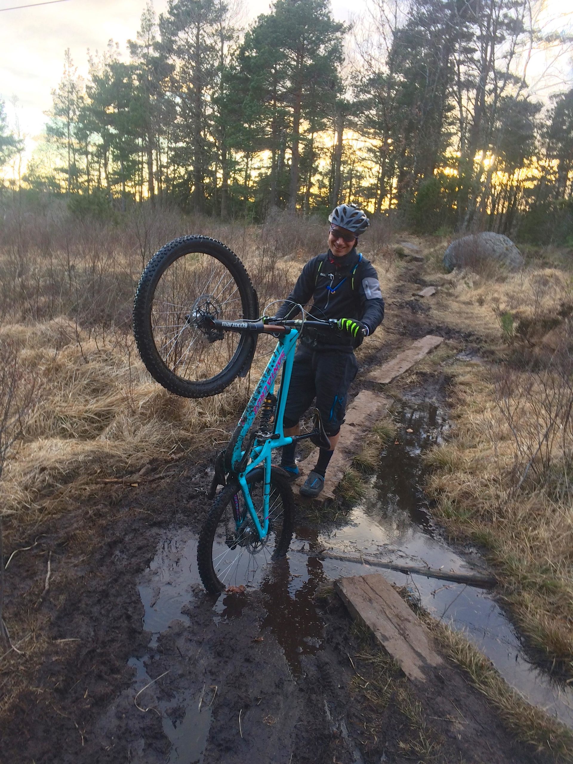 A mountain biker in protective gear carrying their turquoise bike upright while navigating a muddy trail with puddles. The background features trees and a sunset sky. Skatas Trail System mountain bike trail.