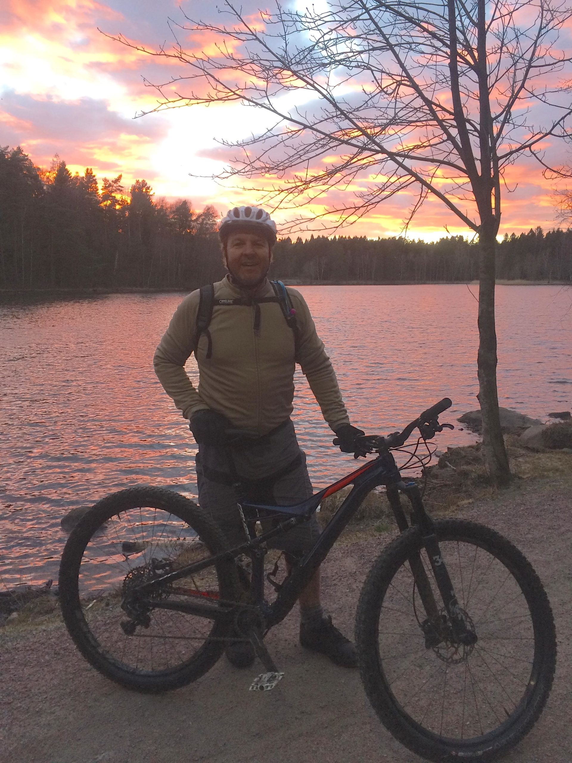 A man in a cycling helmet and gloves stands beside his mountain bike on a path near a lake during sunset. The sky is filled with vibrant shades of pink and orange, and trees line the background, reflecting the warm colors. Skatas Trail System mountain bike trail.