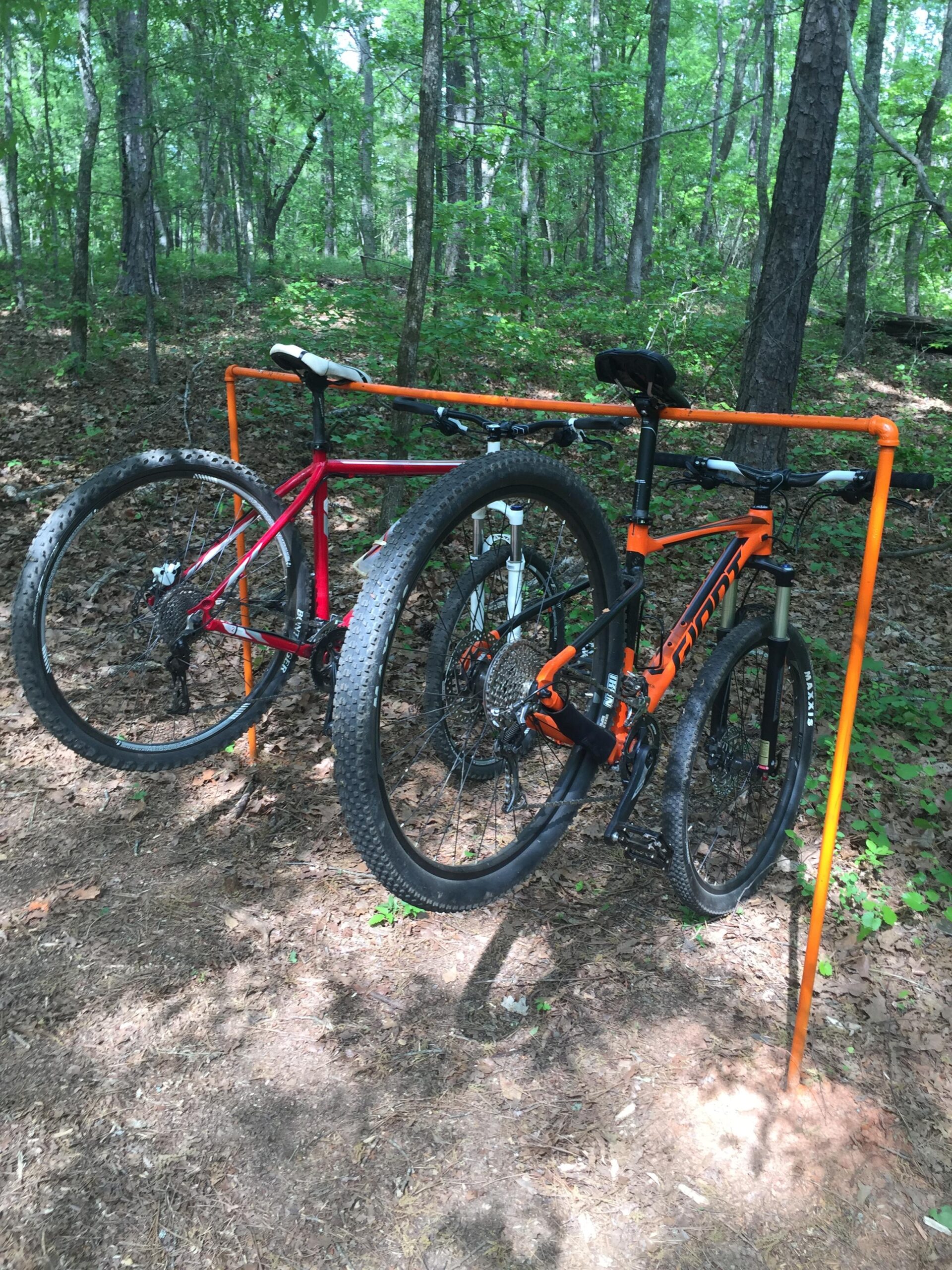 Giant Giant Fathom: Two mountain bikes, one red and one orange, are parked on an orange bike rack in a wooded area. The ground is covered with leaves and dirt, surrounded by lush green foliage and trees. The scene conveys a sense of outdoor adventure and cycling activity.