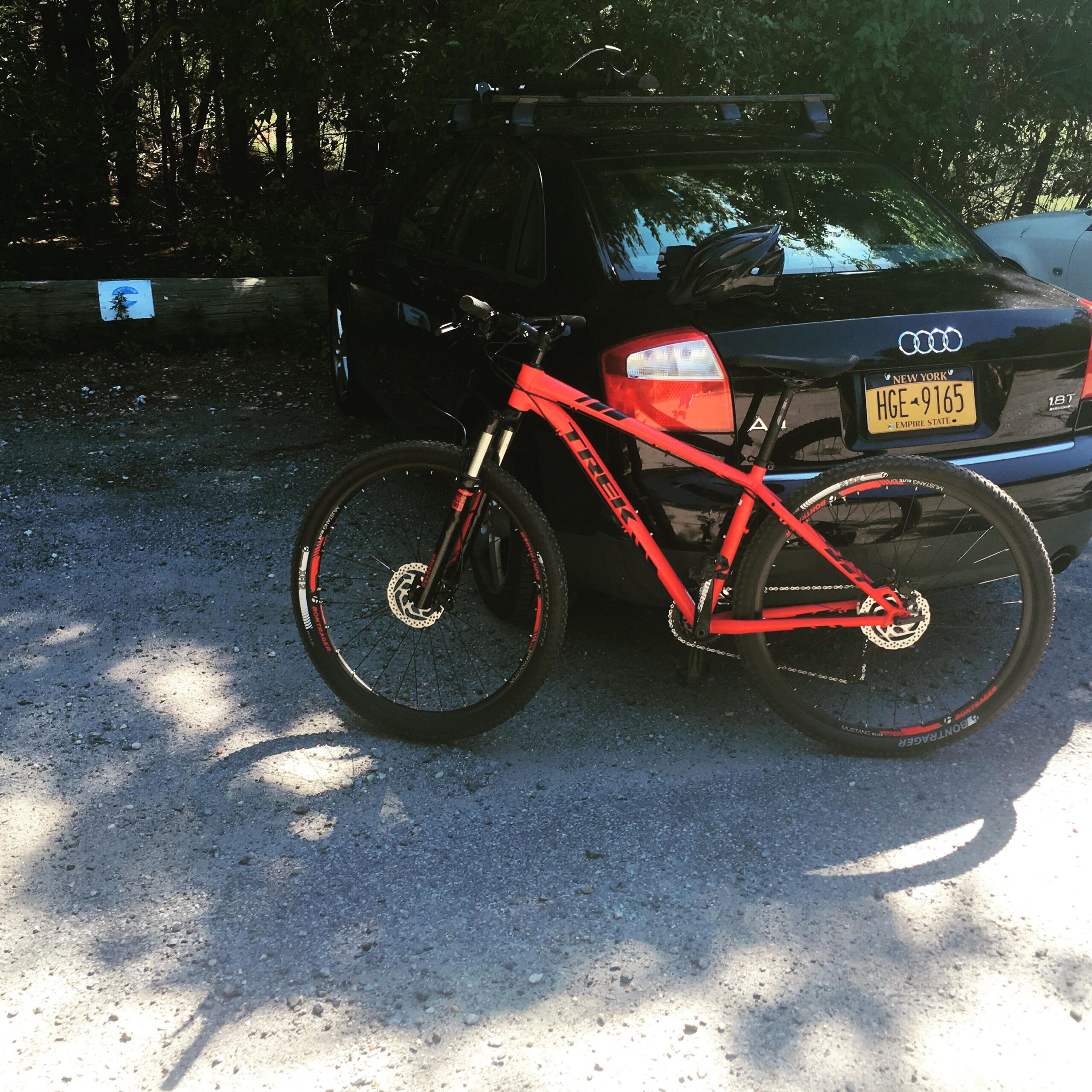 Trek Trek x caliber 8: A red mountain bike leaning against a black car parked on a gravel surface, with trees in the background and a blue sign partially visible in the distance.