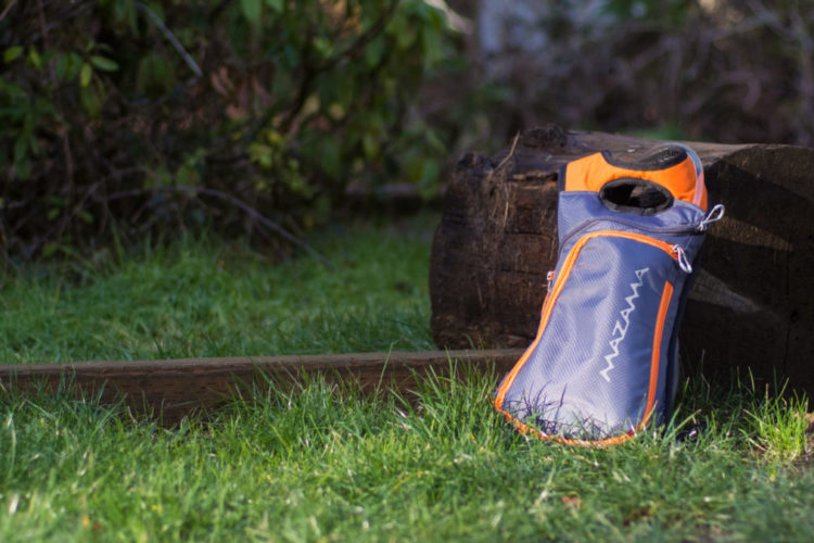 A lightweight, gray and orange outdoor hydration pack resting on lush green grass near a log, surrounded by natural vegetation.