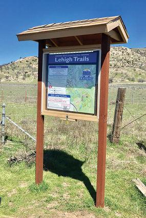 A wooden signpost displaying a map and information about Lehigh Trails, set in a grassy area with hills in the background under a clear blue sky. TMTA Lehigh trails mountain bike trail.