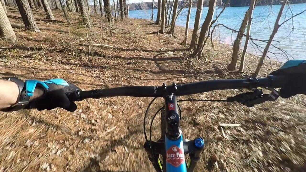 A close-up view of a mountain bike handlebar as it navigates a dirt path lined with trees, with a body of water visible in the background. Autumn leaves cover the ground, and the sun shines through the trees, indicating a warm day for outdoor riding. Liberty Reservior Trails mountain bike trail.