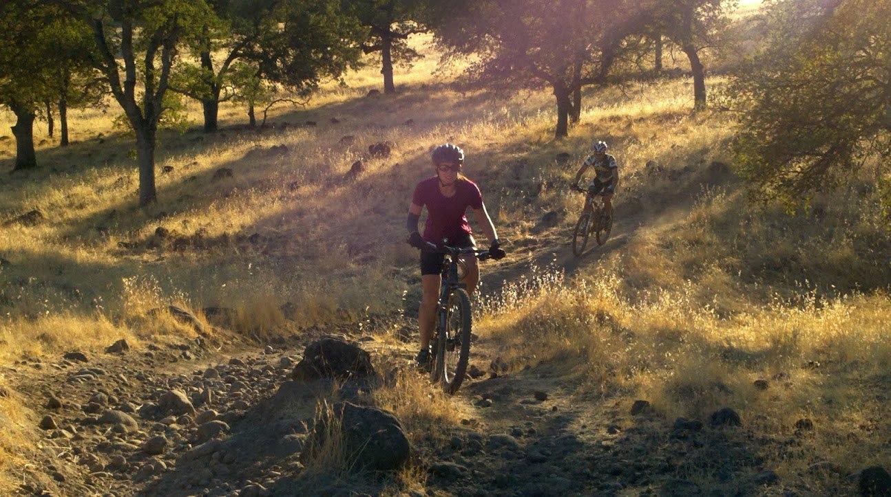 Two mountain bikers riding on a rocky trail surrounded by tall grasses and trees during the golden hour. One cyclist, wearing a red shirt and helmet, is in the foreground, while another cyclist is visible in the background. The sunlight creates a warm, natural glow in the scene. Middle Trail mountain bike trail.