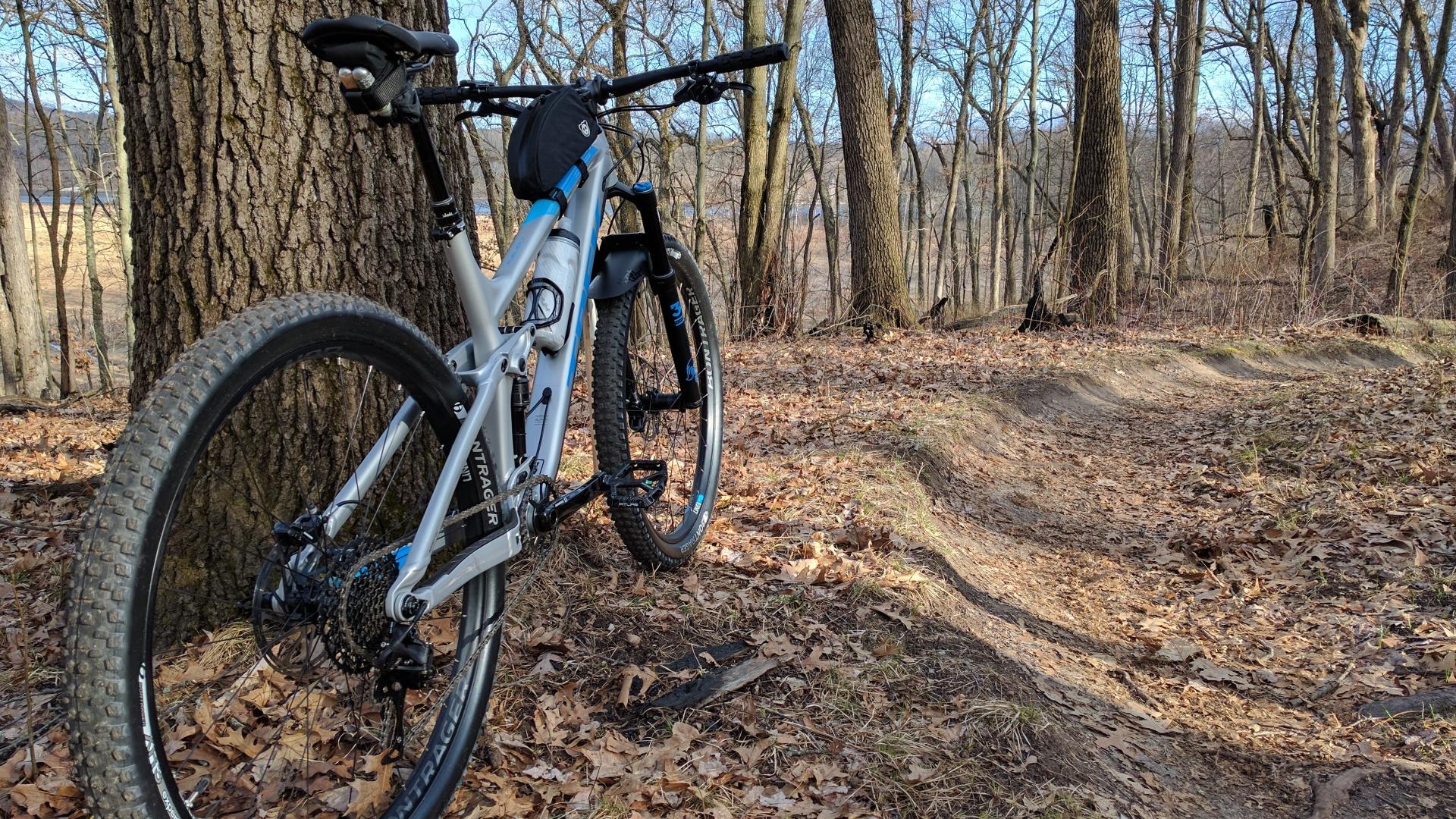 Trek Fuel EX 9 29: A mountain bike leaning against a tree on a trail surrounded by bare trees and fallen leaves. The bike features a blue and gray frame, thick tires, and a small bag mounted on the handlebars. A winding dirt path can be seen in the background.