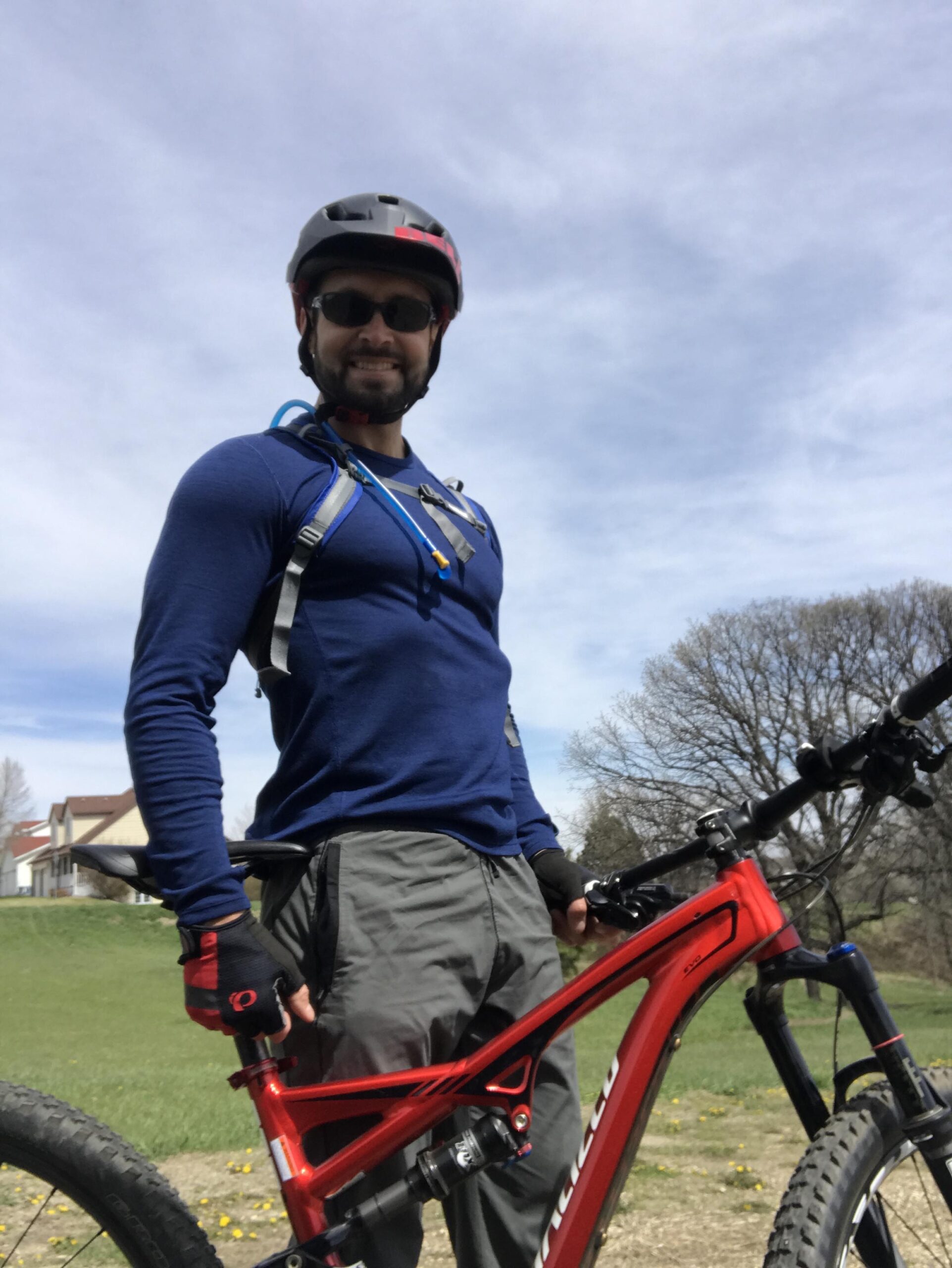 Specialized Camber 29: A smiling male cyclist wearing a helmet and sunglasses stands next to a red mountain bike. He is dressed in a blue long-sleeve shirt and gray pants, with gloves on his hands. The background features green grass and a clear sky with a few clouds.