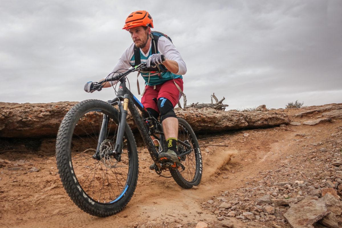 A cyclist riding a mountain bike on a sandy, rugged trail, navigating around a large rock formation. The rider is wearing a helmet, gloves, and knee pads, and is dressed in a short-sleeved shirt and shorts. The cloudy sky adds a dramatic backdrop to the scene, emphasizing the outdoor adventure. Big Mesa mountain bike trail.