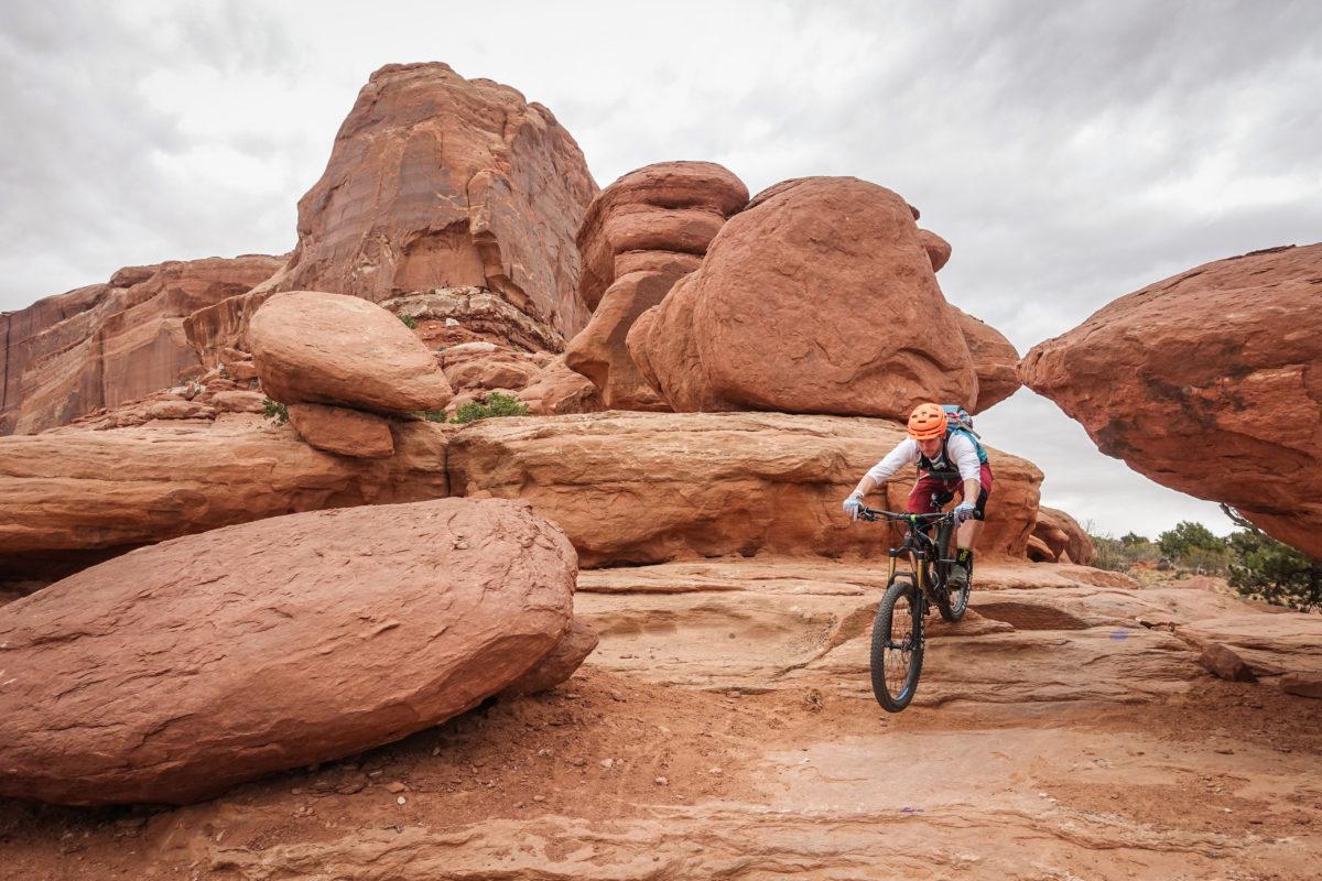 A mountain biker navigating through a rocky landscape with large sandstone formations, under an overcast sky. The rider is wearing an orange helmet and a backpack, focused on maneuvering over rough terrain. Big Mesa mountain bike trail.