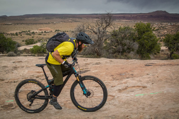 A mountain biker in a yellow shirt and helmet rides on rocky terrain, leaning forward as he navigates the trail. He is equipped with a backpack and gloves, and the landscape features distant hills and sparse vegetation under a cloudy sky.