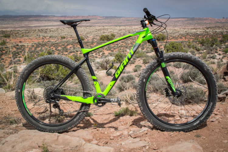 A green and black mountain bike is positioned on a rocky surface with a vast desert landscape in the background. The bike features thick tires and a lightweight frame, emphasizing its designed for off-road performance. The scene captures a rugged environment with sparse vegetation and distant hills under a cloudy sky.