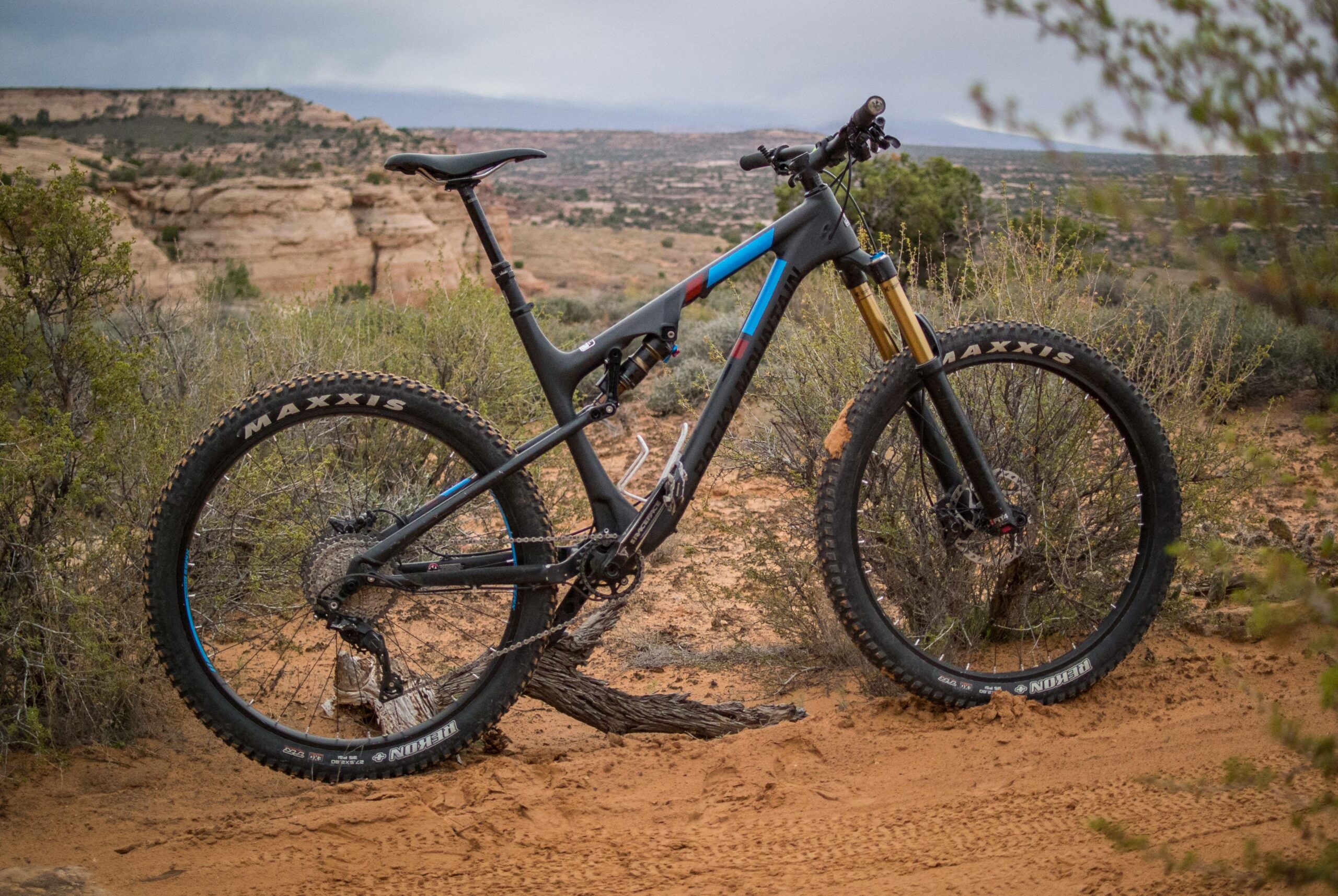 Rocky Mountain Pipeline: A black and blue mountain bike leaning against a small tree in a sandy desert landscape, with rocky formations and sparse vegetation in the background. The bike features large tires with "Maxxis" branding and is positioned on a patch of sandy ground.
