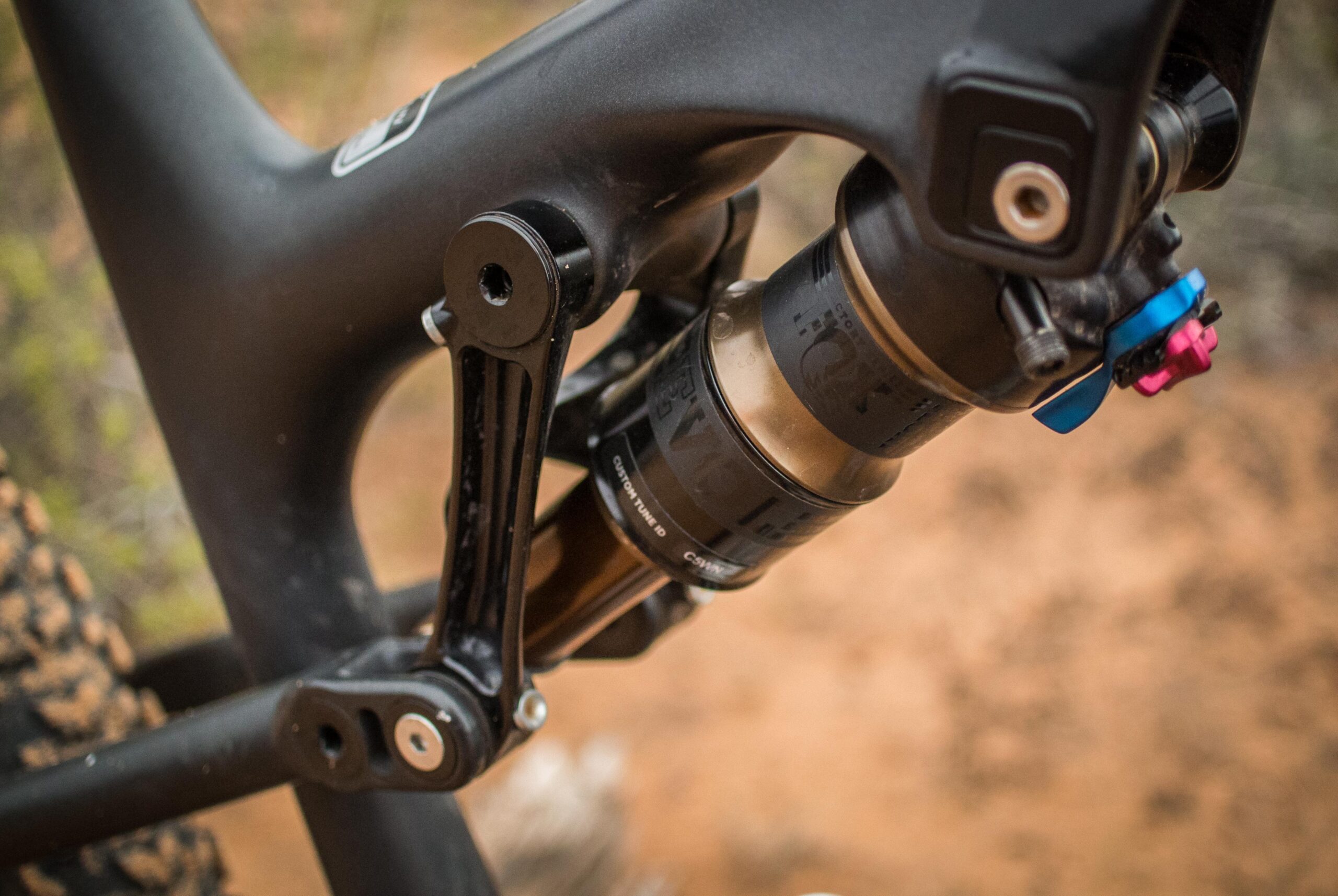 Rocky Mountain Pipeline: Close-up view of a bicycle's rear suspension system, featuring a black frame with a shock absorber and adjustable tuning options. The background shows a blurred natural setting, likely a trail or outdoor area.