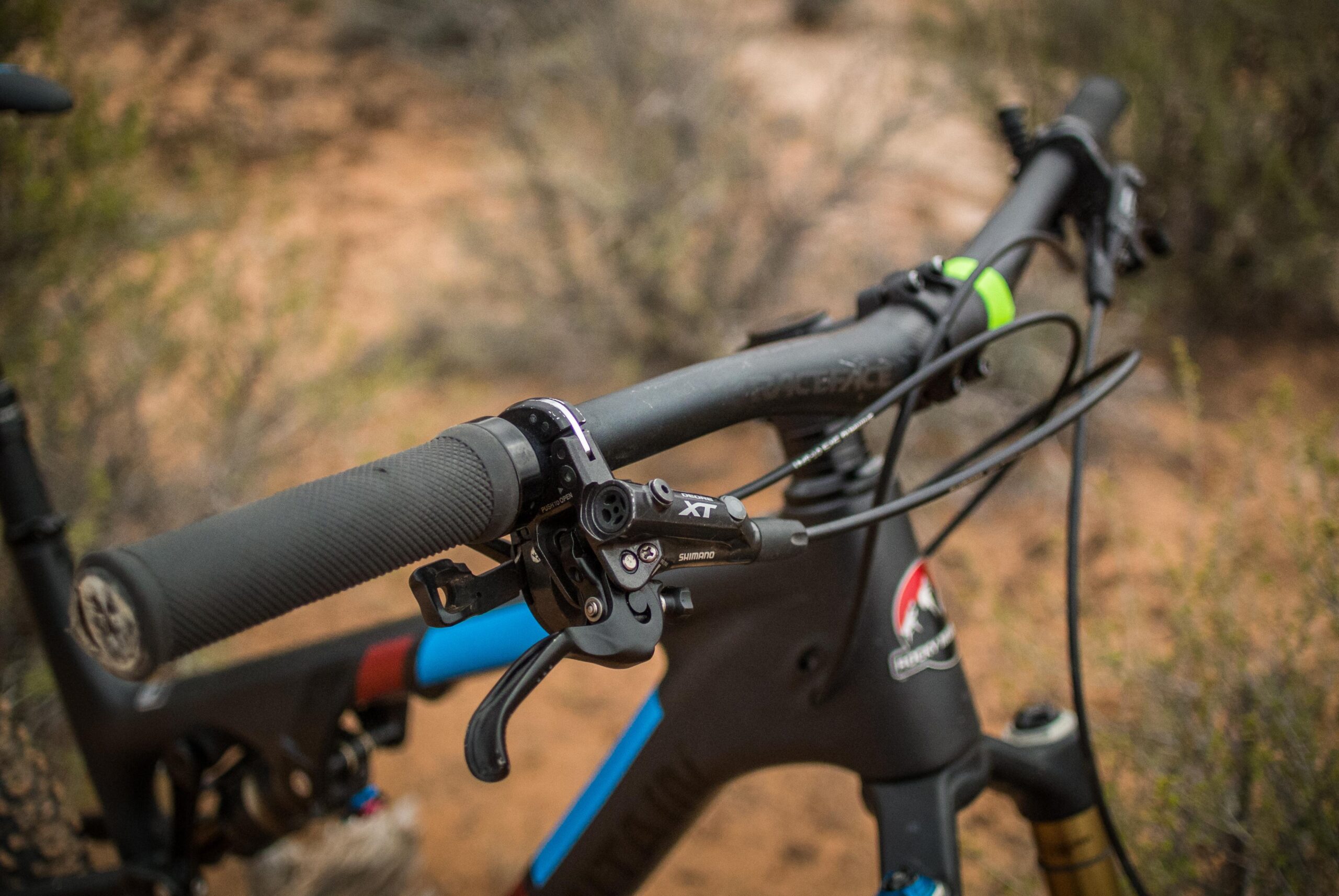 Rocky Mountain Pipeline: Close-up view of a mountain bike handlebar with textured black grips, hydraulic brake lever, and gear shifter, set against a blurred natural background.