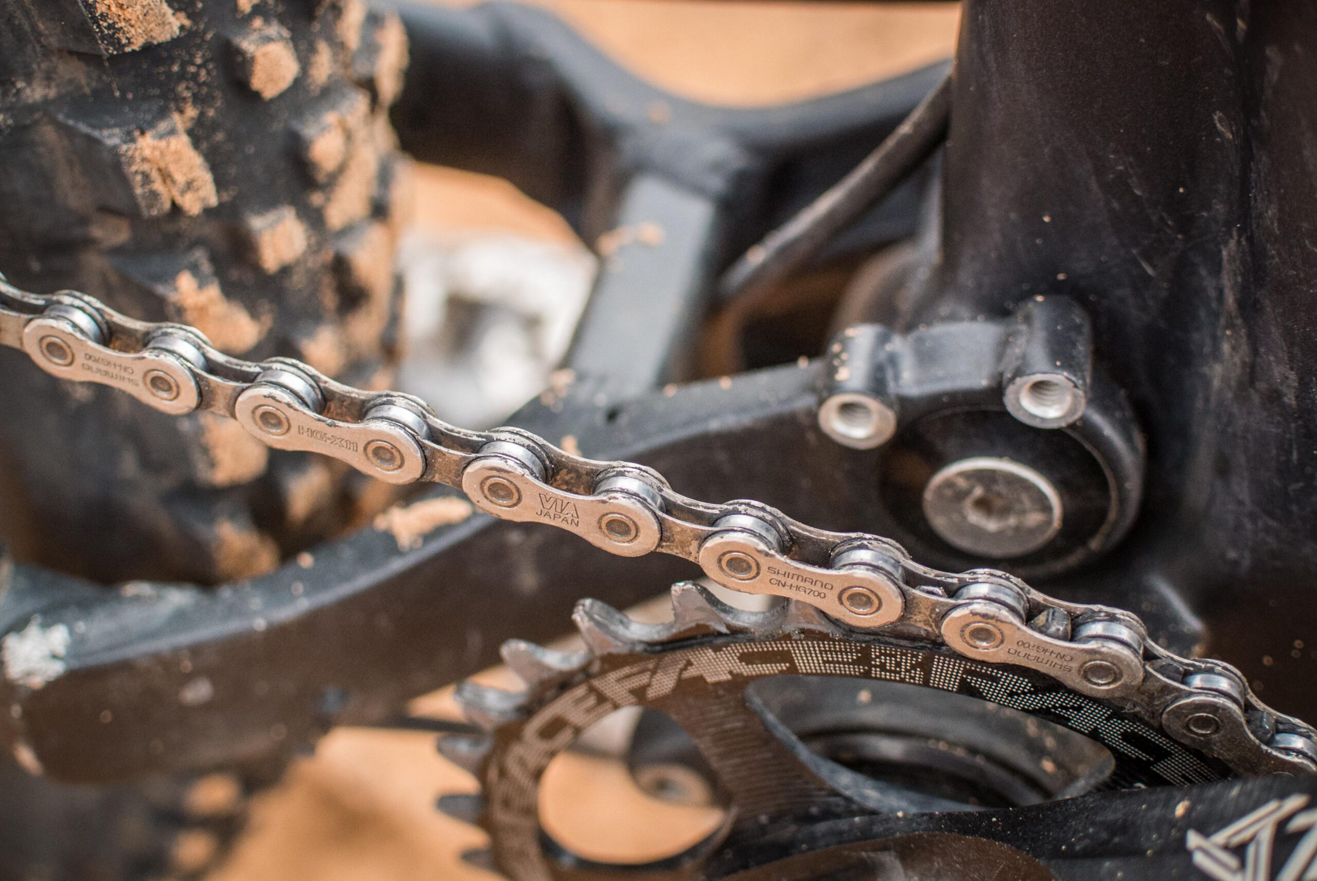 Rocky Mountain Pipeline: Close-up view of a bicycle chain and gears, showing intricate details of the chain links and a black bike frame partially covered in dirt. The image highlights the mechanical components and the wear from usage.