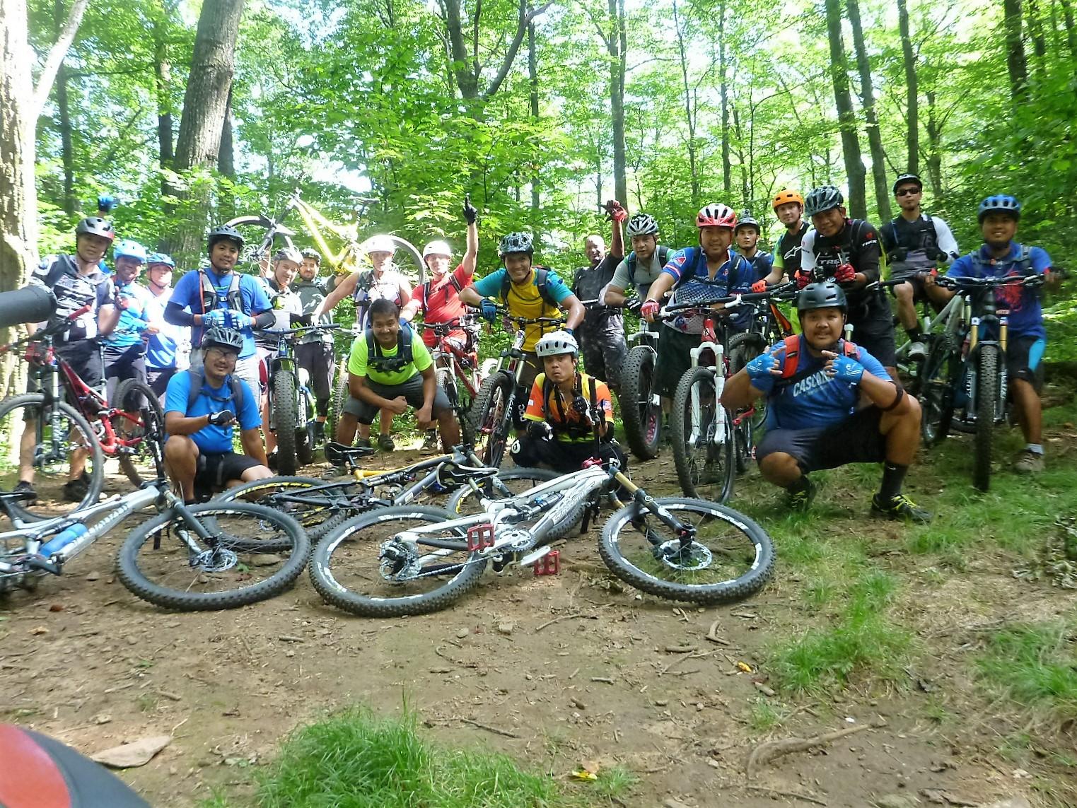 A group of mountain bikers posing together in a wooded area, surrounded by trees. Some riders are crouching in front, while others stand behind them, holding their bikes. The scene is vibrant, showcasing various colorful clothing and helmets, with several bicycles lying in the foreground. Pilot Rock mountain bike trail.