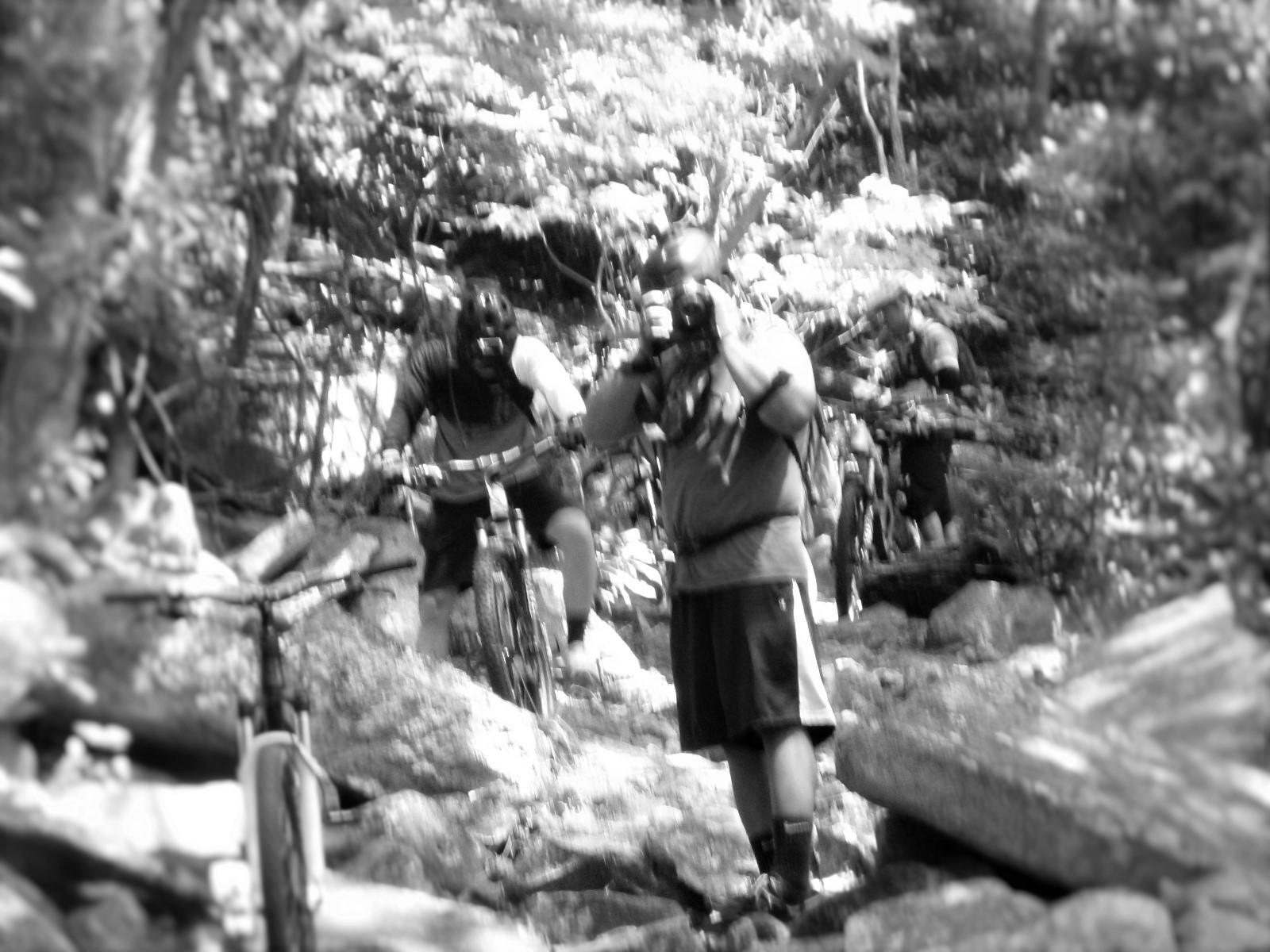 A group of mountain bikers navigating a rocky trail in a forest, with one rider pausing to take a photo. The image is in black and white, highlighting the surrounding nature and the rugged terrain. Pilot Rock mountain bike trail.