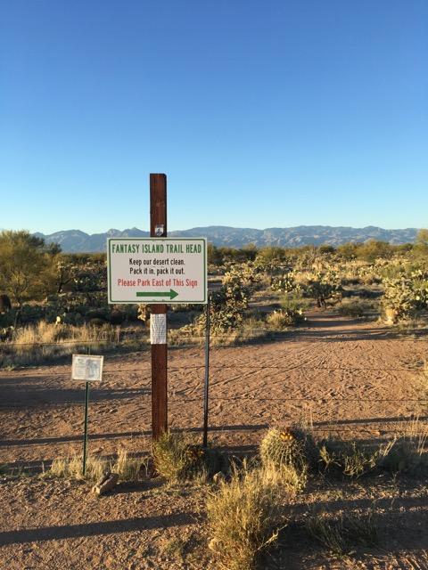 A dirt trail leading towards a mountainous landscape with a signpost directing visitors to "Fantasy Island Trail Head." The sign emphasizes keeping the area clean and indicates parking should be done to the east of the sign. Surrounding vegetation includes desert plants and cacti under a clear blue sky. Fantasy Island mountain bike trail.