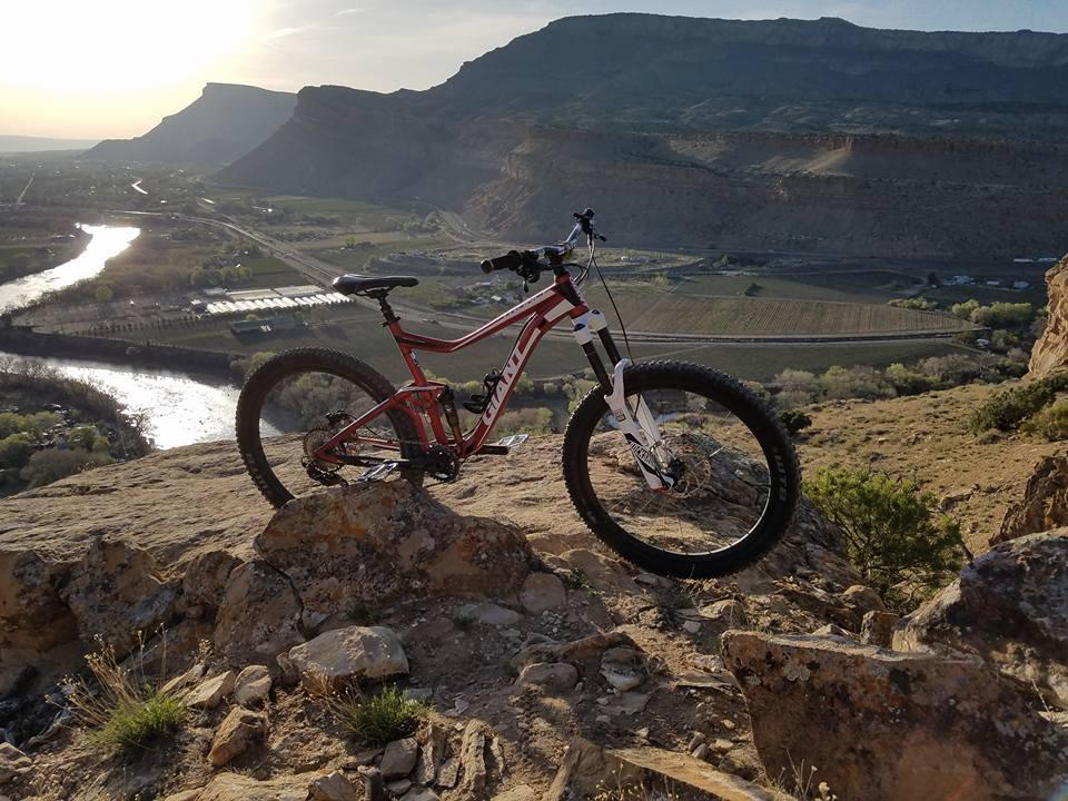Giant Trance 27.5 3: A red mountain bike positioned on rocky terrain overlooking a scenic valley with a river and fields in the background. The landscape features mountains in the distance and a clear sky.
