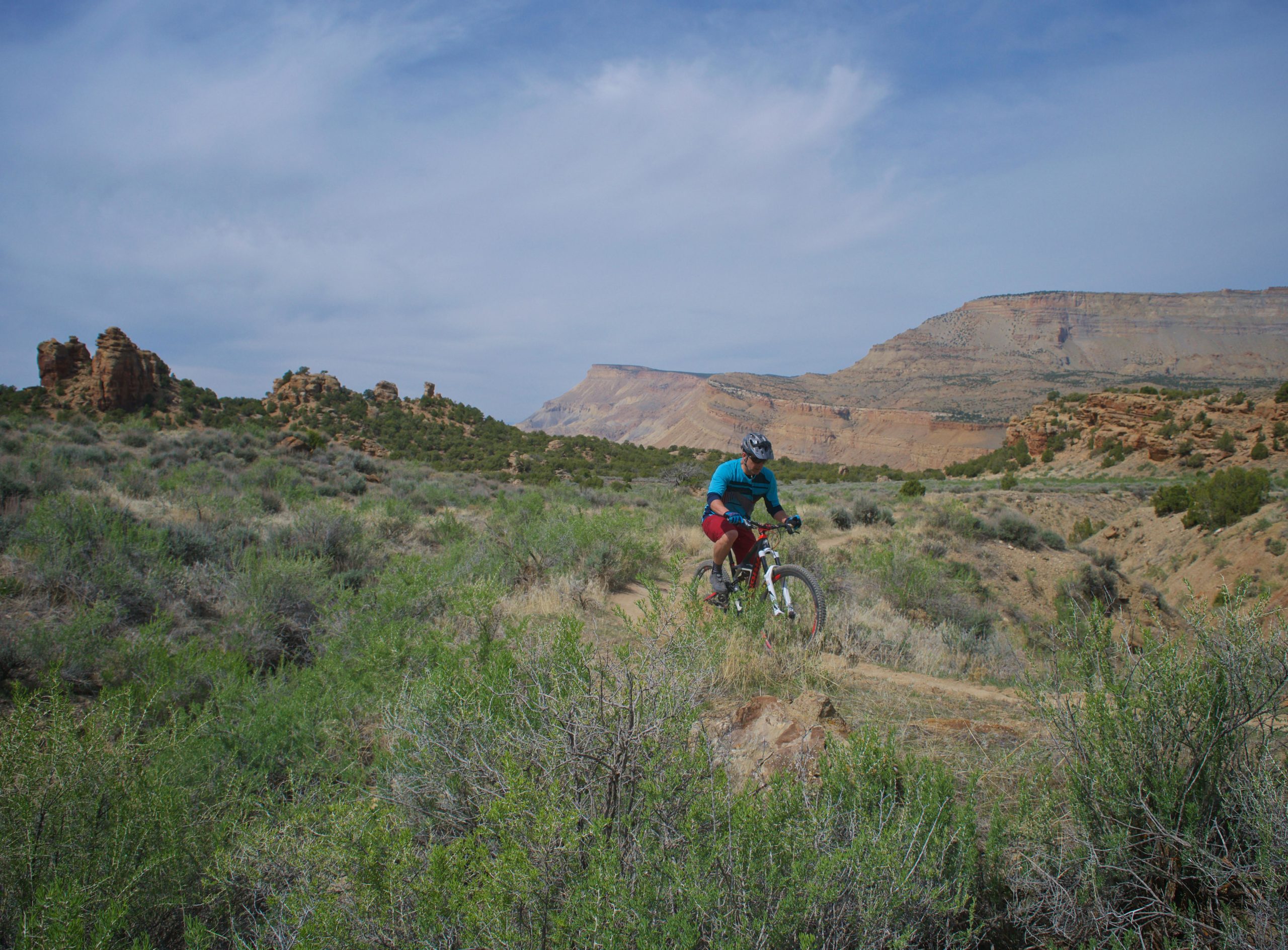 A mountain biker rides through a rugged, desert landscape with rocky formations and rolling hills in the background. The scene features sparse vegetation and a clear blue sky, capturing the essence of outdoor adventure. Palisade Rim mountain bike trail.