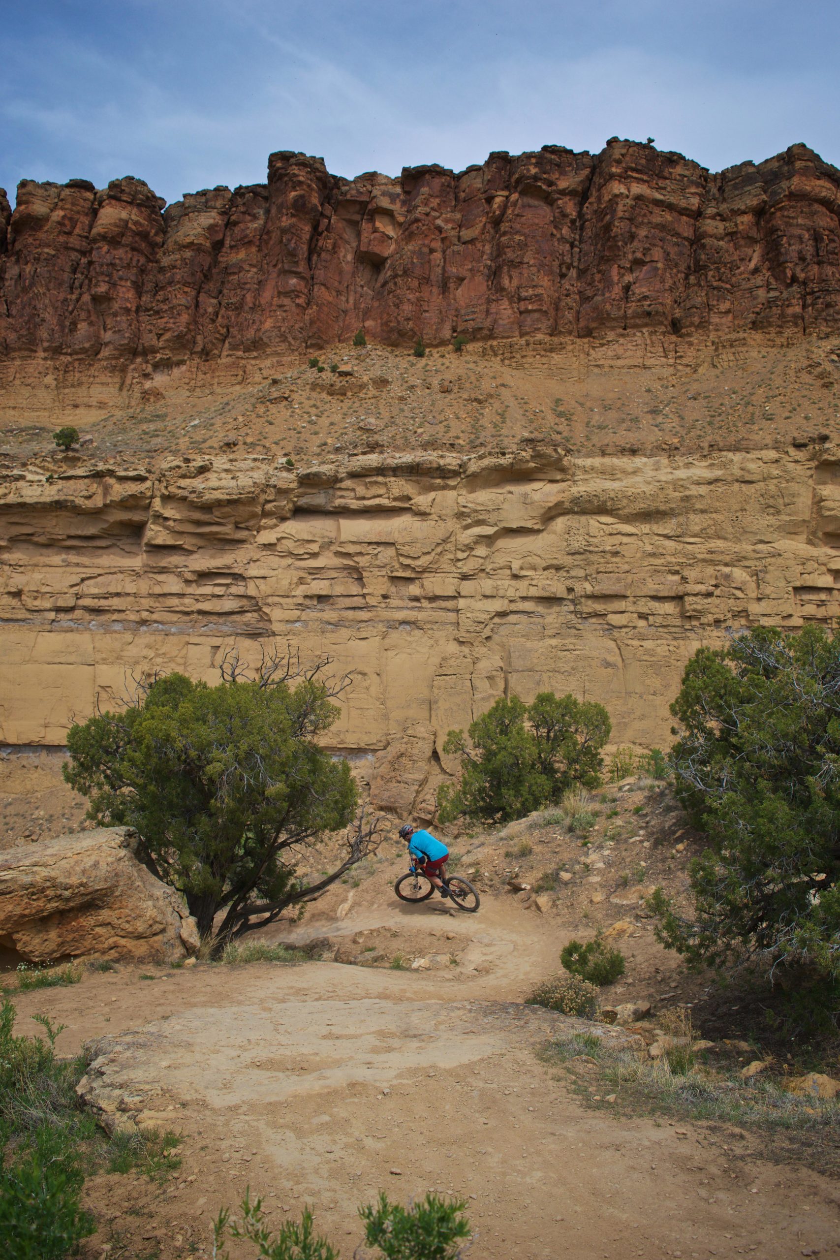 A mountain biker in a blue shirt navigates a dirt trail in a rocky landscape, surrounded by steep, reddish cliffs and sparse vegetation. The sky is partly cloudy. Palisade Rim mountain bike trail.