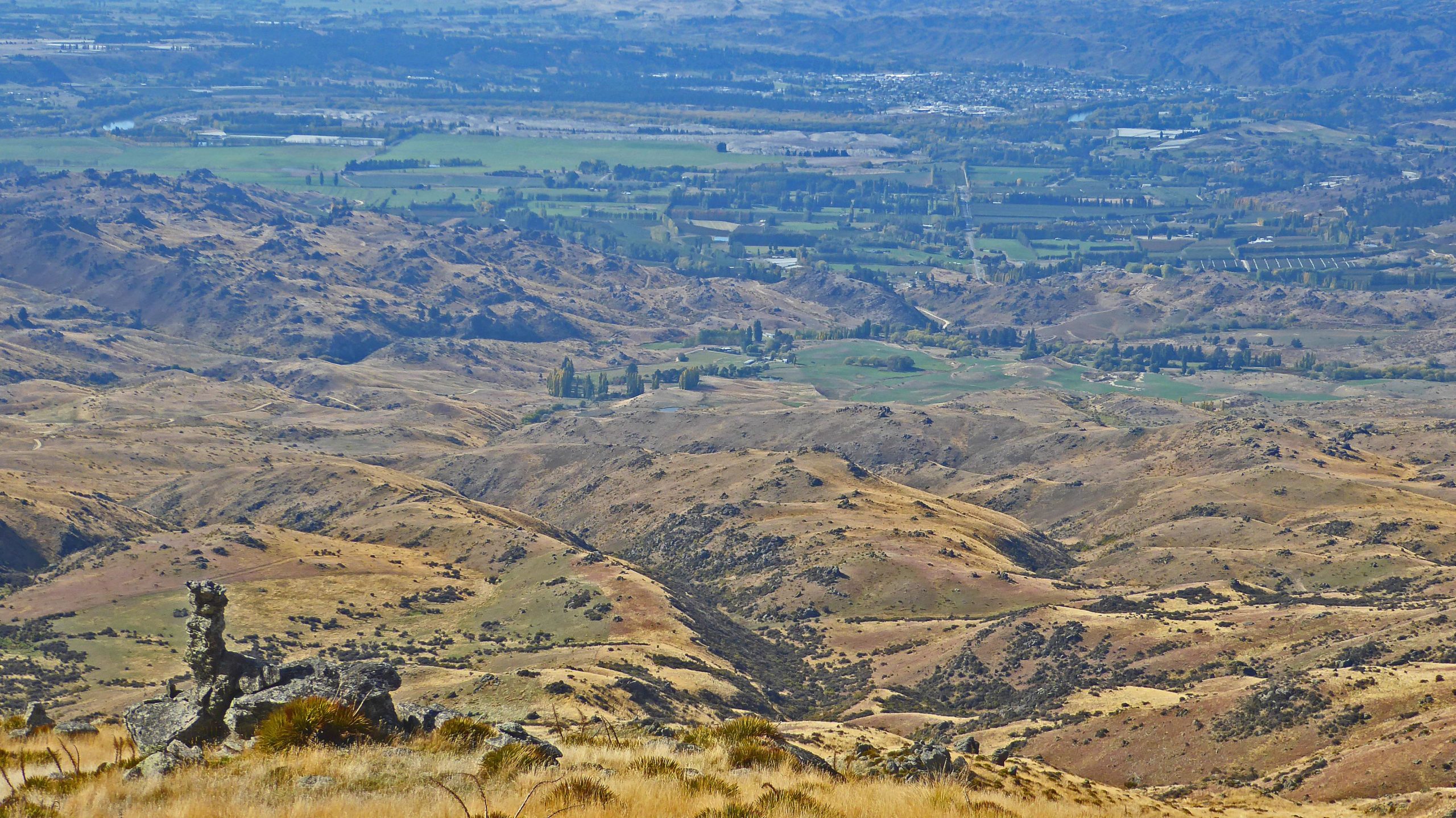 A panoramic view of rolling hills and valleys, featuring a mix of dry grassland and patches of green fields. In the foreground, a rocky outcrop is visible, while the background reveals a broader landscape with distant mountains and farmland. The scene is captured under a clear blue sky, highlighting the natural beauty of the countryside. Obelisk mountain bike trail.
