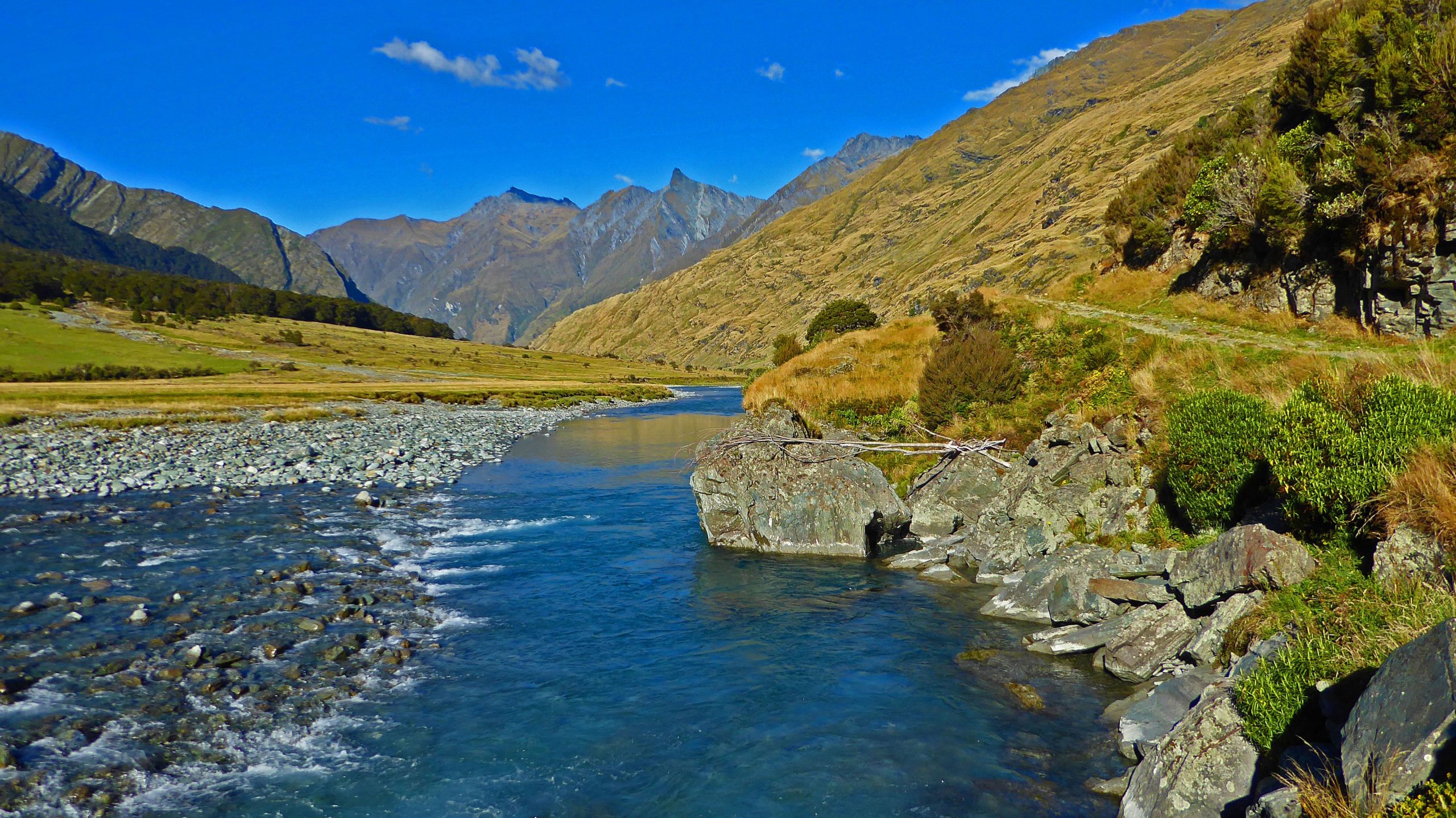 A scenic view of a river winding through a valley, surrounded by rolling hills and mountains under a clear blue sky. The river features rocky banks, and the landscape includes patches of greenery and dry grass. Aspiring Hut mountain bike trail.