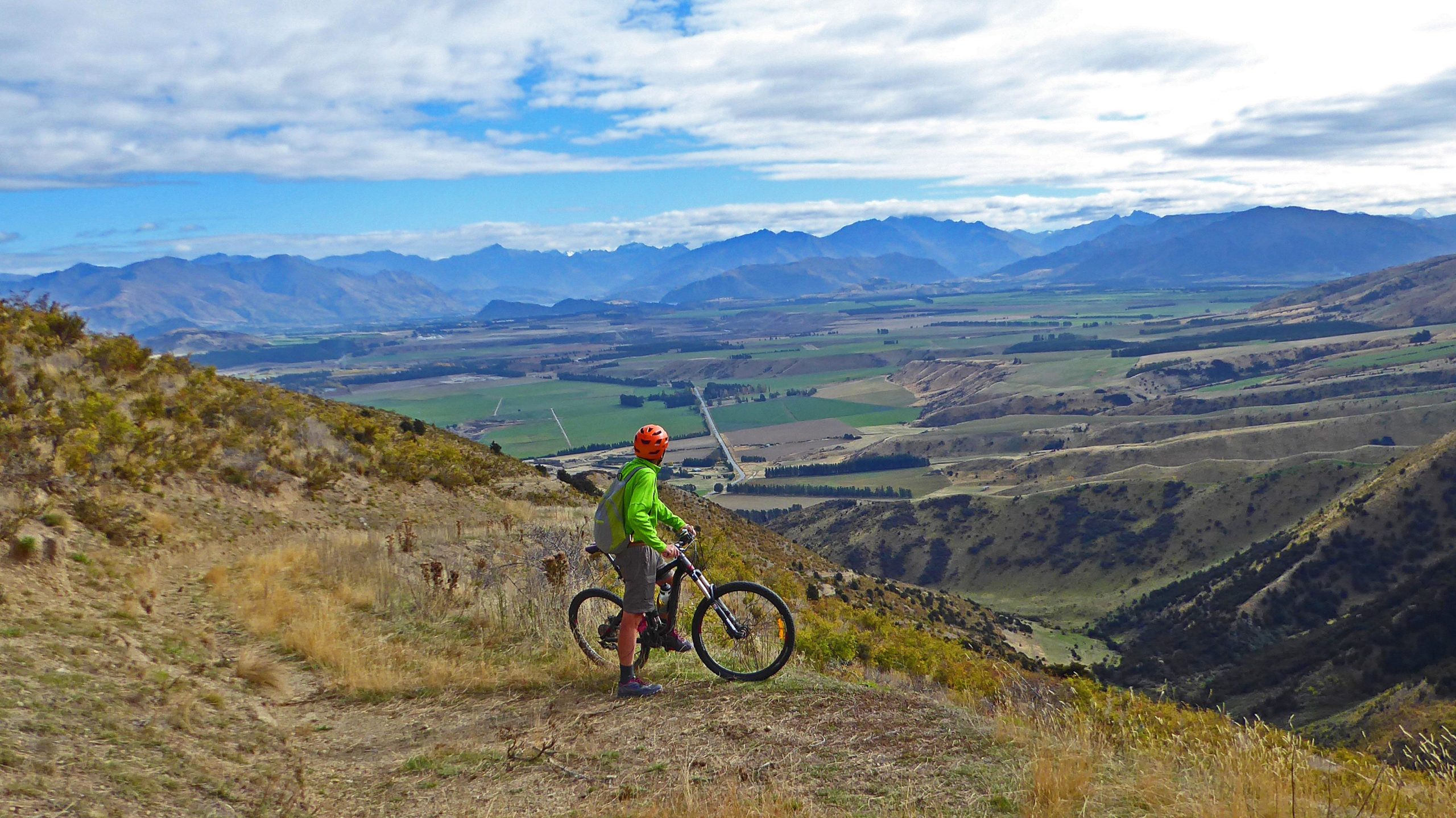 A mountain biker in a bright green jacket and orange helmet stands on a hillside, overlooking a lush valley and distant mountain ranges under a partly cloudy sky. The terrain is a mix of grass and shrubs, with winding roads and fields visible in the landscape below. Grandview Ridge Track mountain bike trail.