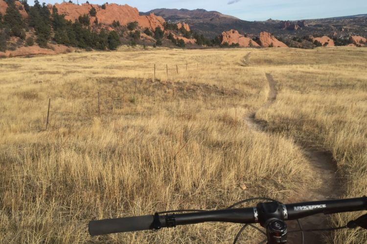 A view from the handlebars of a mountain bike, overlooking a grassy field with tall, dry grass and a dirt path winding through it. In the background, red rock formations rise against a clear sky, indicating a natural landscape, possibly in a mountainous area.