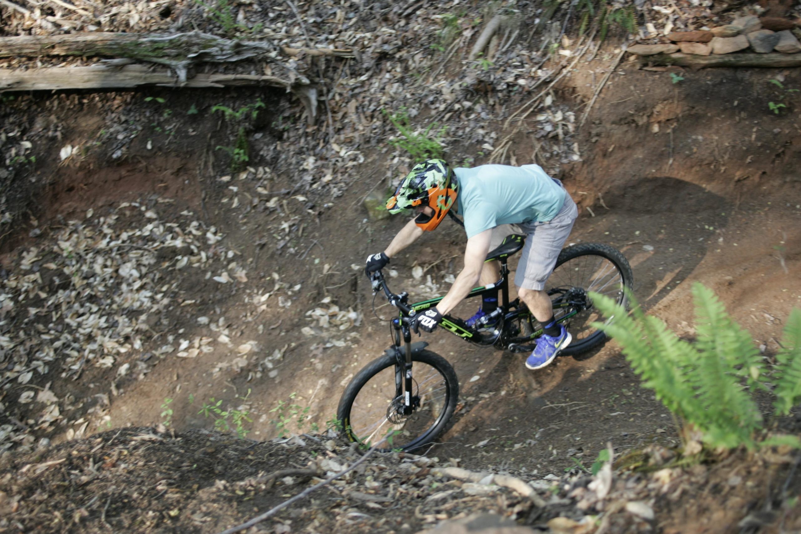 A person riding a mountain bike down a dirt trail surrounded by foliage and leaf litter, wearing a helmet and gloves for safety. The biker leans forward, showing dynamic movement as they navigate the terrain. Salem Lake mountain bike trail.