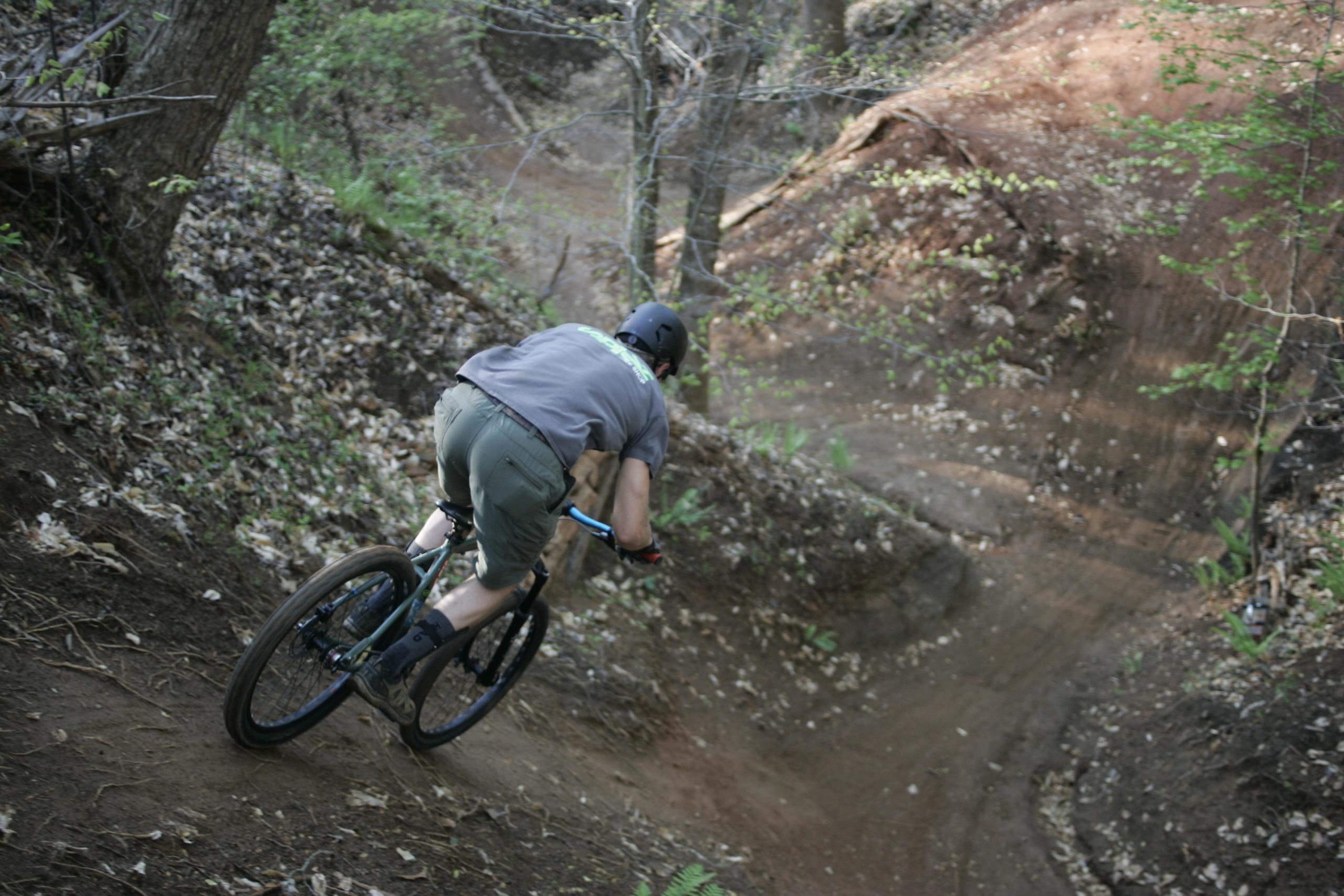 A mountain biker riding down a dirt trail surrounded by trees and leaves, leaning into a turn on a rugged, natural terrain. Salem Lake mountain bike trail.