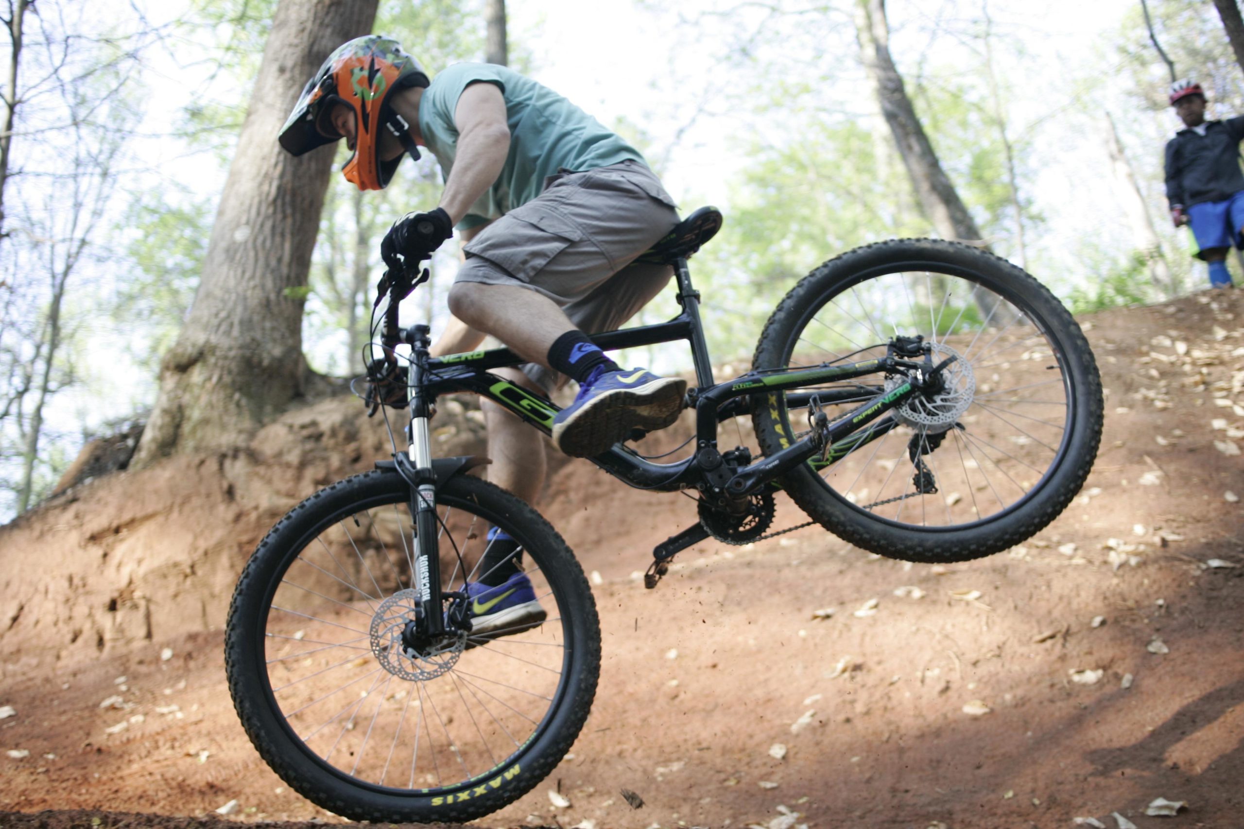A person riding a mountain bike is mid-jump on a dirt trail surrounded by trees. The cyclist is wearing a helmet and gloves, and is positioned in a dynamic stance, with one foot off the pedal. The background shows a dirt ramp and forest setting. Salem Lake mountain bike trail.