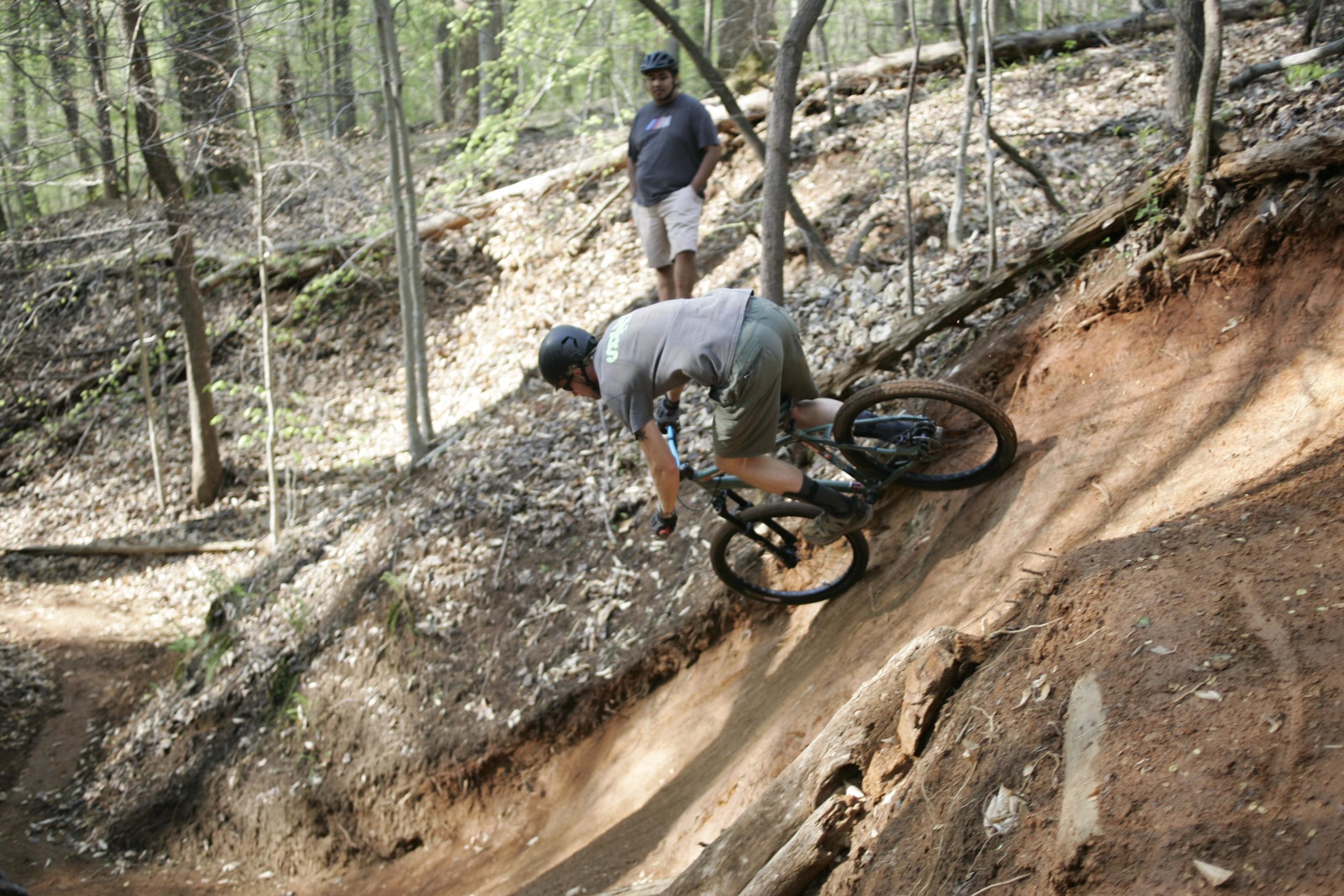 A mountain biker skillfully navigates a dirt trail in a wooded area, leaning into a turn. In the background, another person watches from the side, surrounded by trees and fallen leaves. Salem Lake mountain bike trail.